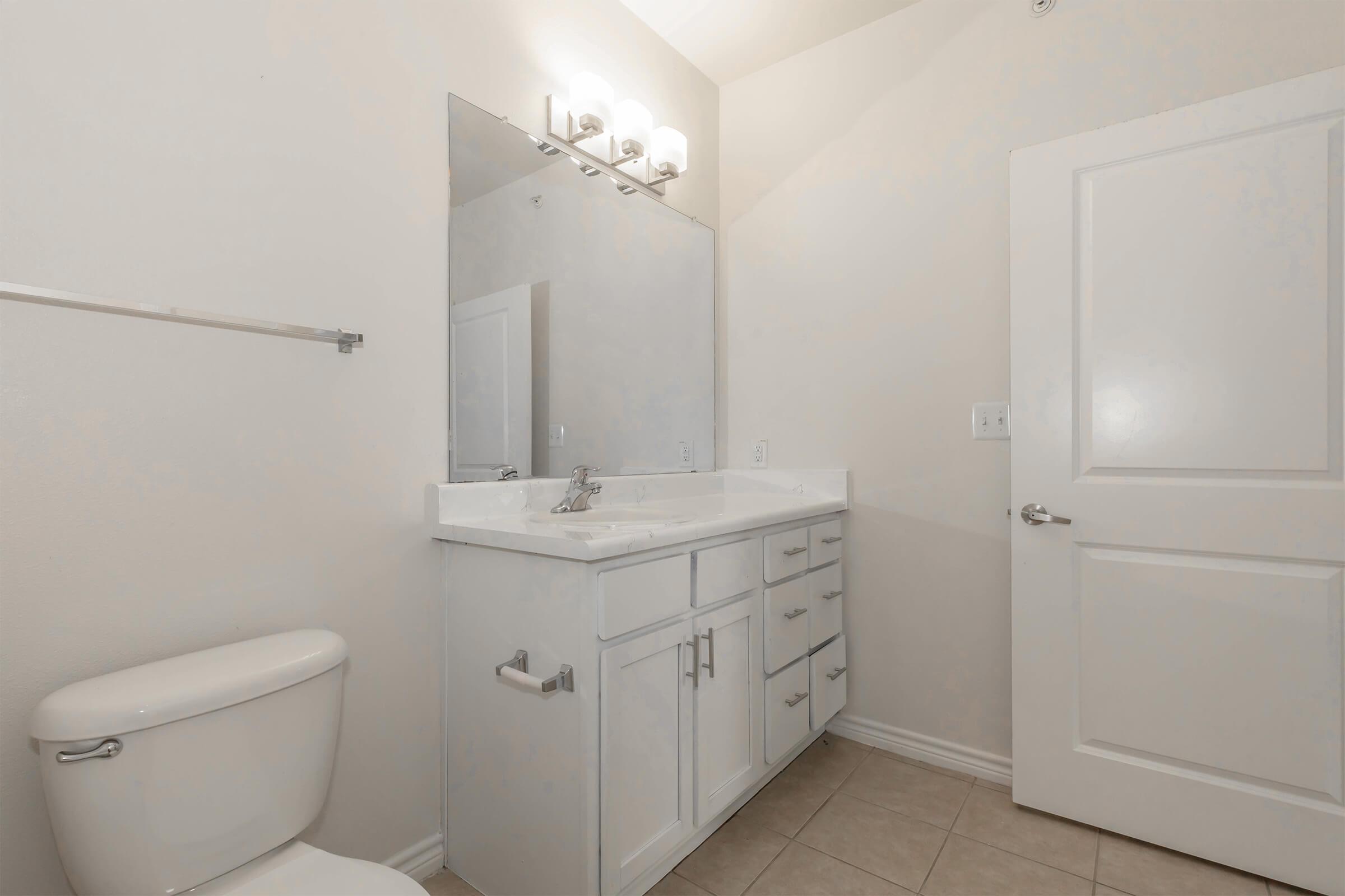 A clean and modern bathroom featuring a white toilet, a double sink vanity with a marble countertop, and a large mirror above it. The walls are painted light colors, and there are light fixtures above the mirror, enhancing the bright and airy feel of the space. A door leads to another room.
