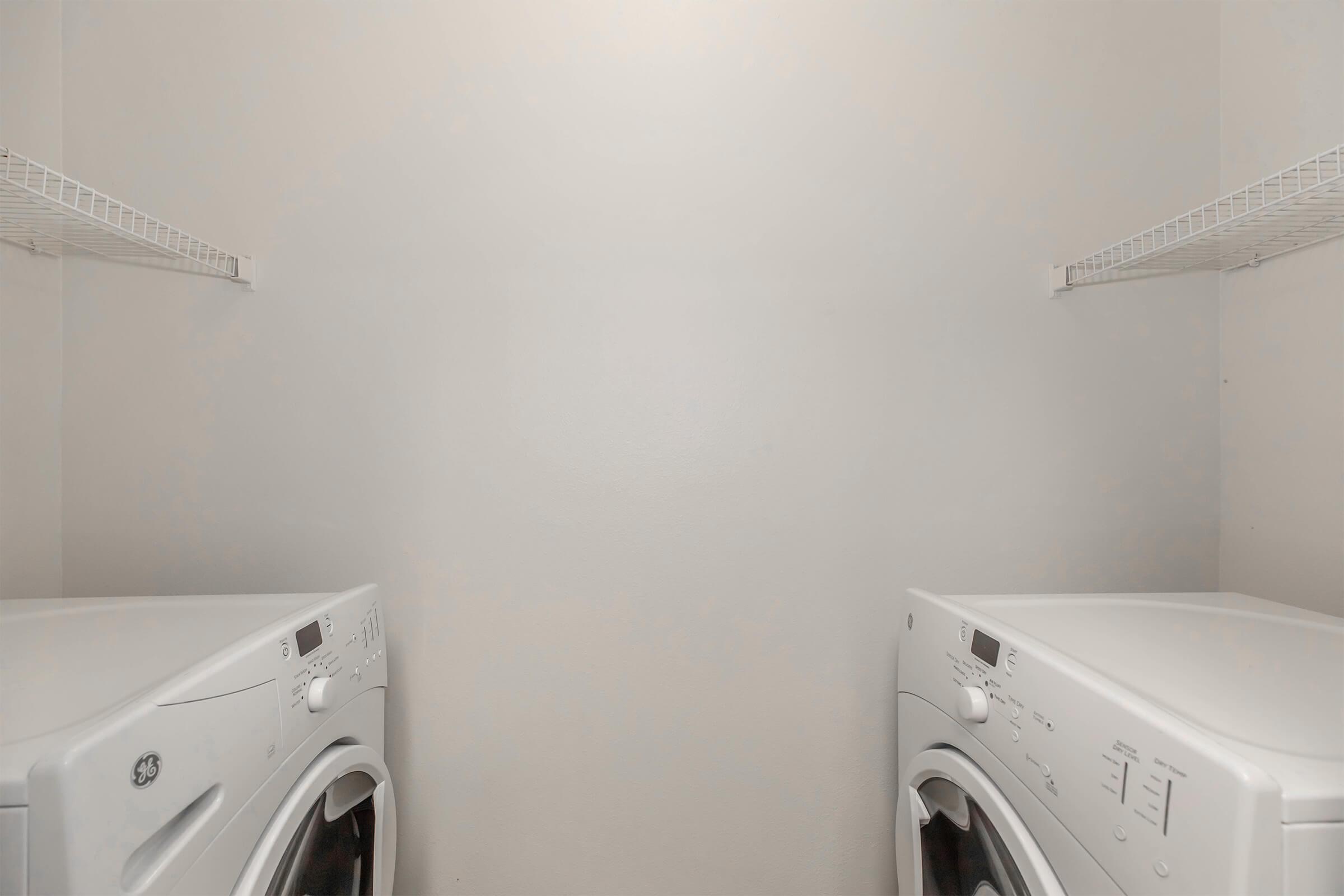 A small laundry room with two white washing machines side by side. The walls are light-colored, and there are wire shelves above the machines for storage. The space appears empty and well-lit, with a simple and clean aesthetic.