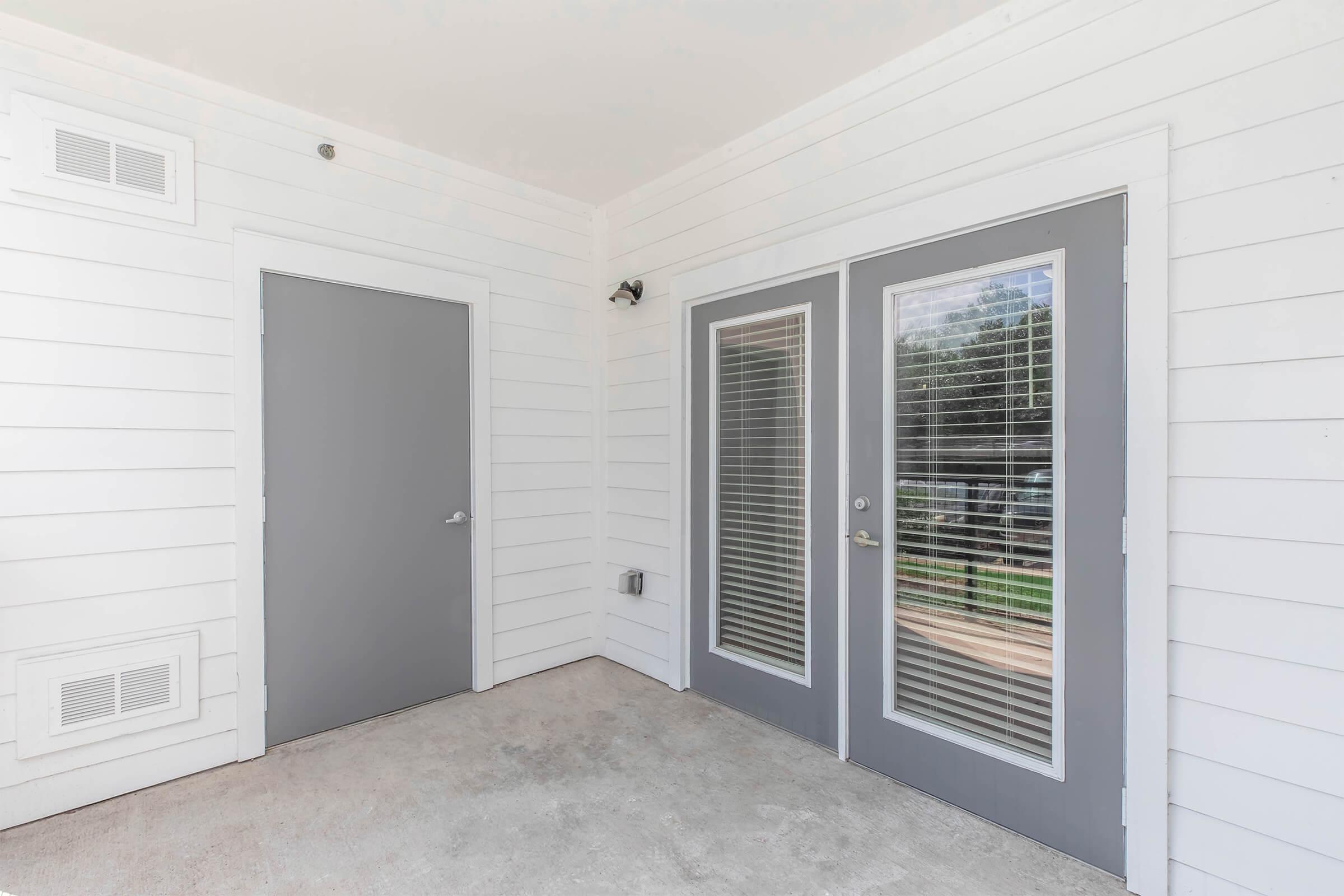 Exterior view of a small porch area featuring three doors: one solid gray door and two glass-paneled doors with gray frames. The walls are white shiplap, and there's a light fixture above the doors. The floor is concrete, and there are windows visible outside.