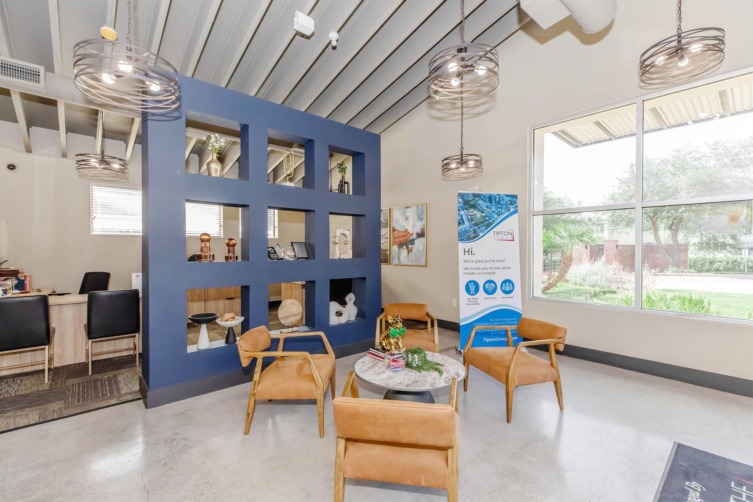 A modern indoor lounge area featuring stylish brown chairs around a small table, with a decorative display wall on one side. A banner is visible, and large windows allow natural light into the space, providing a view of greenery outside. The decor is contemporary and inviting.