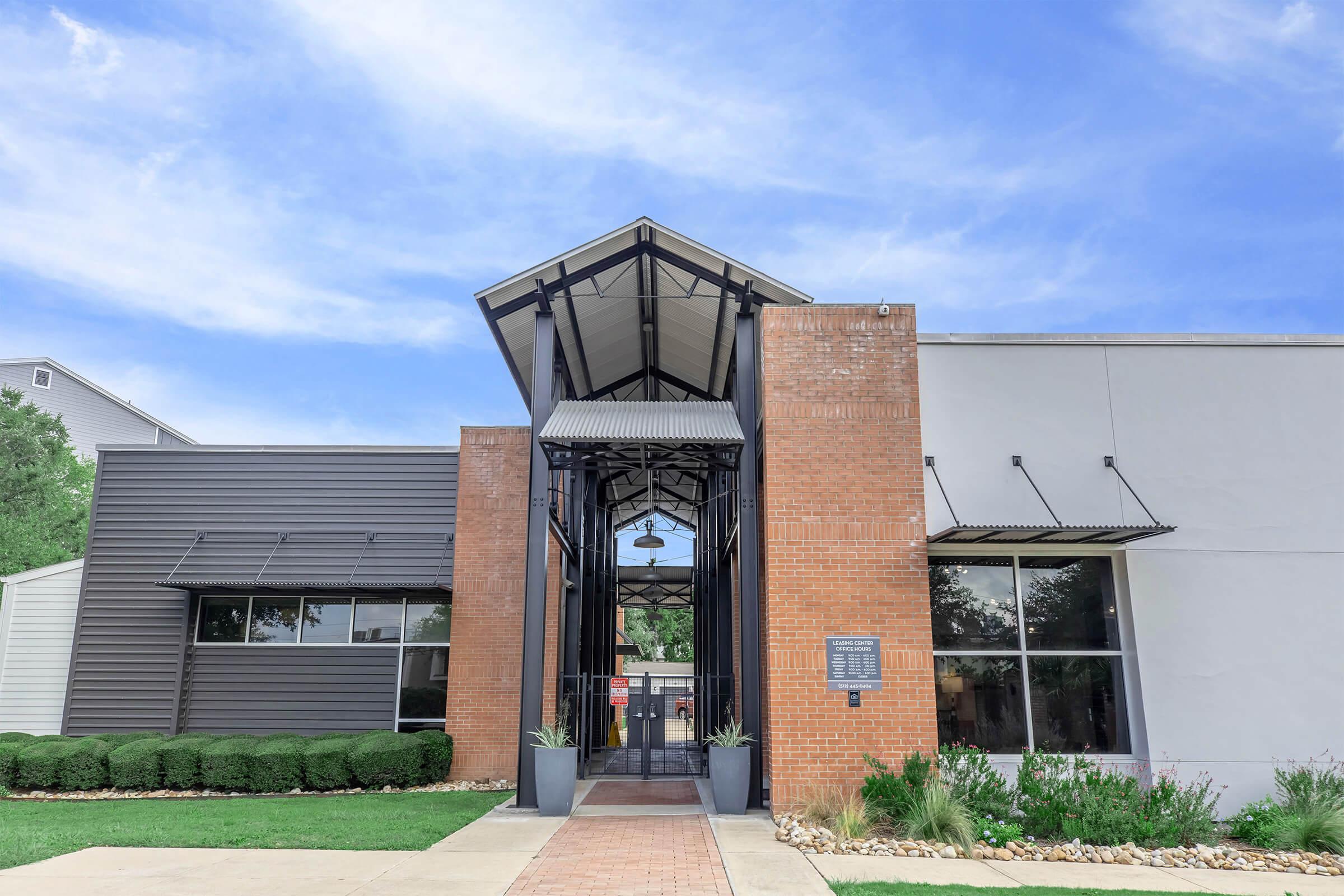 Modern building entrance with a sleek design featuring a gabled roof and large glass doors. The facade combines brick and metal elements, flanked by neatly trimmed hedges and a pathway leading to the entrance. Clear blue sky provides a bright backdrop.
