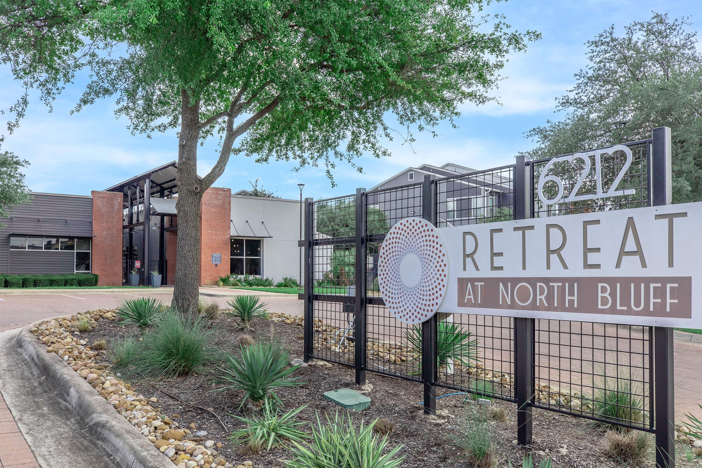 A modern apartment complex entrance featuring a sign that reads "Retreat at North Bluff," with landscaped greenery and stone pathways. The building has a contemporary design with a mix of brick and siding, surrounded by well-maintained grass and shrubs.