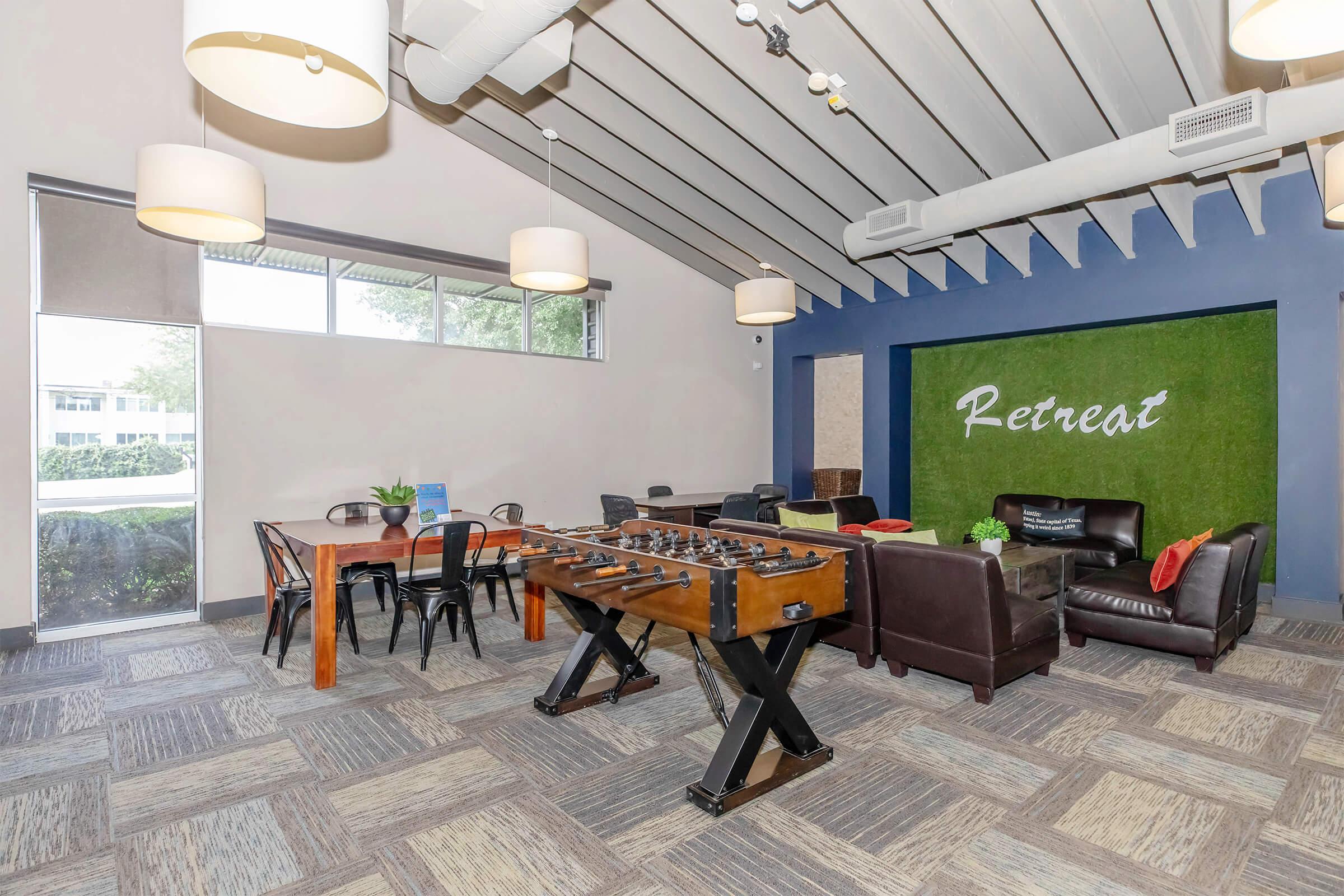 A modern lounge area featuring a foosball table, a large wooden dining table with black chairs, and comfortable seating arrangements against a green "Retreat" wall. The space has large windows allowing natural light, with stylish pendant lighting hanging from a vaulted ceiling.