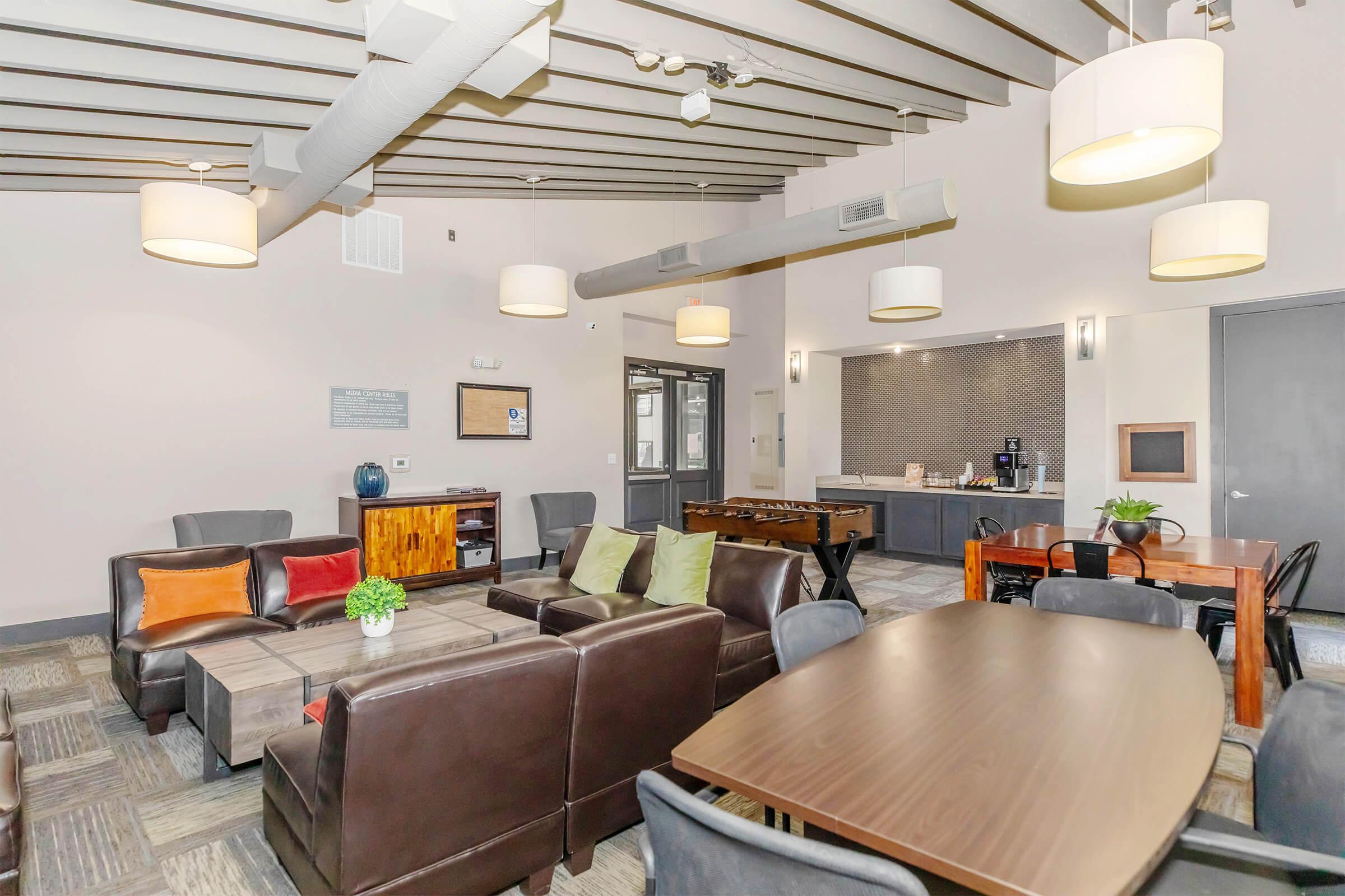 A modern lounge area featuring brown leather sofas with colorful accent pillows, a wooden coffee table, and a dining table. The space has overhead lighting, a gray wall with a coffee station to the right, and textured flooring. Large windows allow natural light to enter, creating a welcoming atmosphere for socializing.