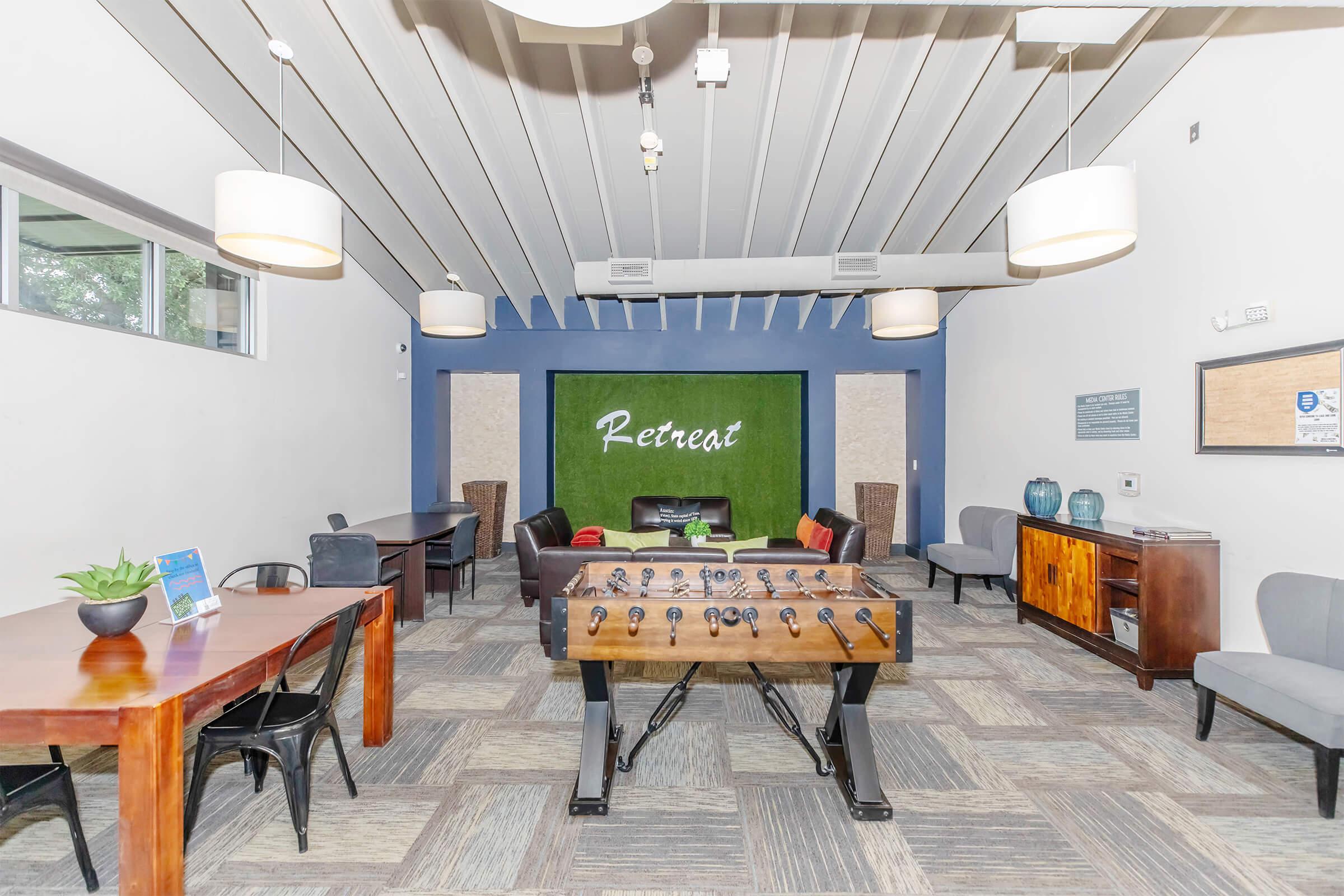 A spacious lounge area featuring a foosball table in the center, surrounded by seating options including chairs and tables. The walls are decorated with green accents, and the word "Retreat" is displayed prominently on the back wall. Soft lighting from hanging lamps enhances the inviting atmosphere.