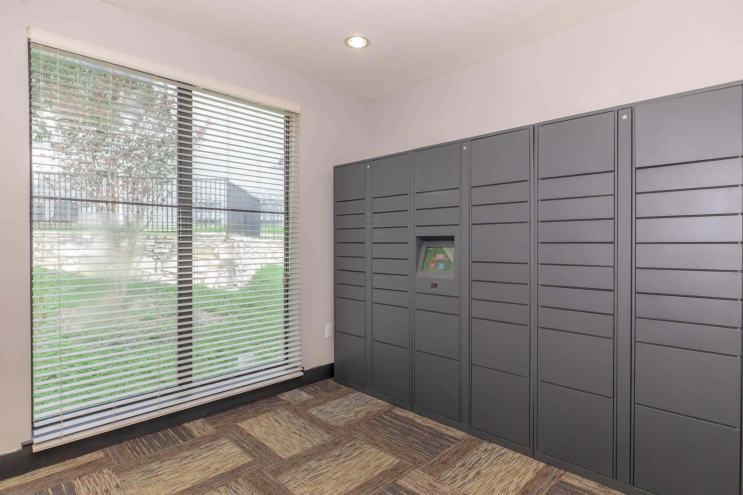 A modern package delivery locker area featuring a row of gray lockers against a wall. A window with horizontal blinds allows natural light to enter the space, illuminating the carpeted floor. The environment appears clean and organized, suitable for residents or users accessing their packages.