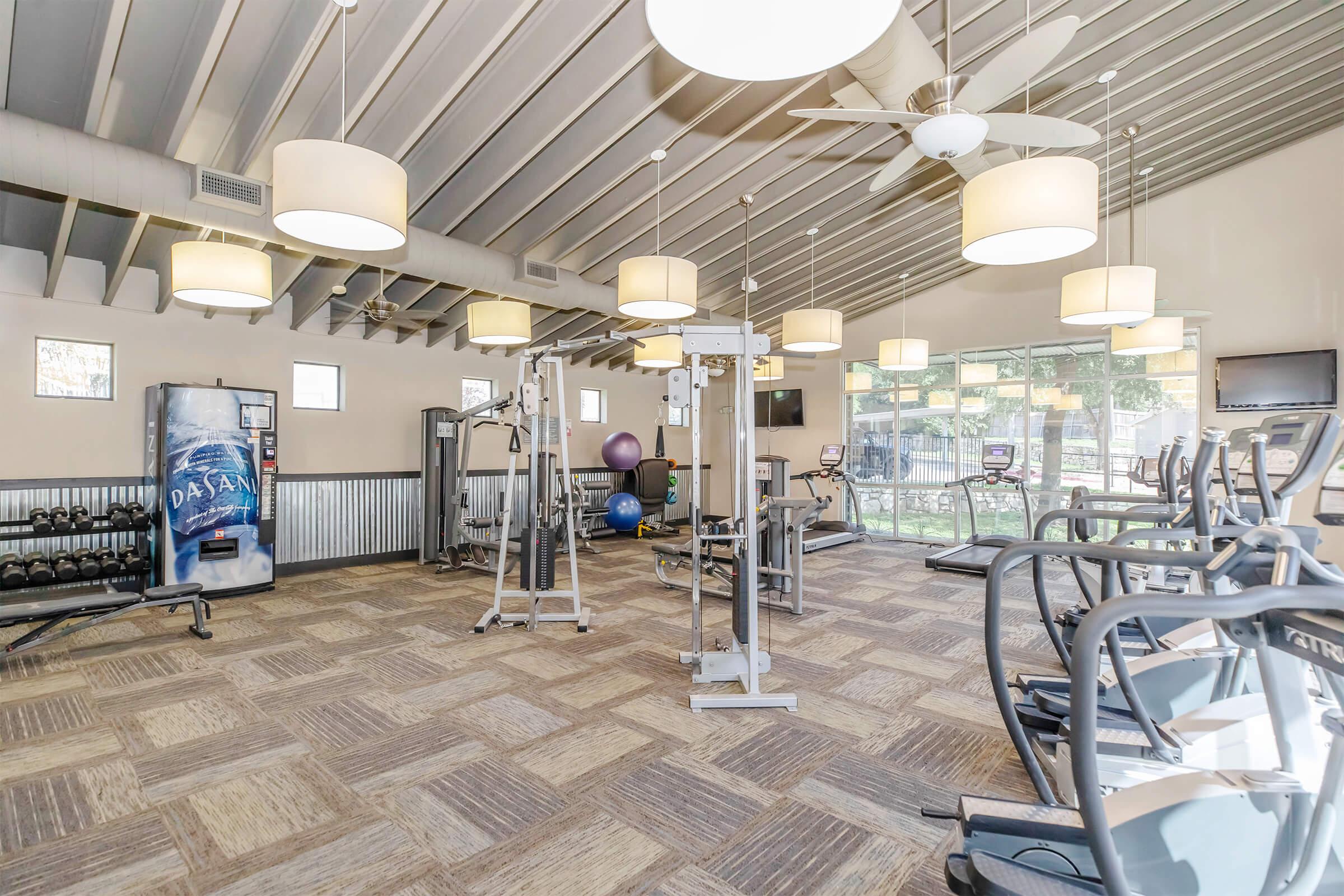 A modern gym interior featuring various exercise equipment, including treadmills, weights, and exercise balls. The space is well-lit with hanging lights and has large windows allowing natural light. A water vending machine is visible in the corner.