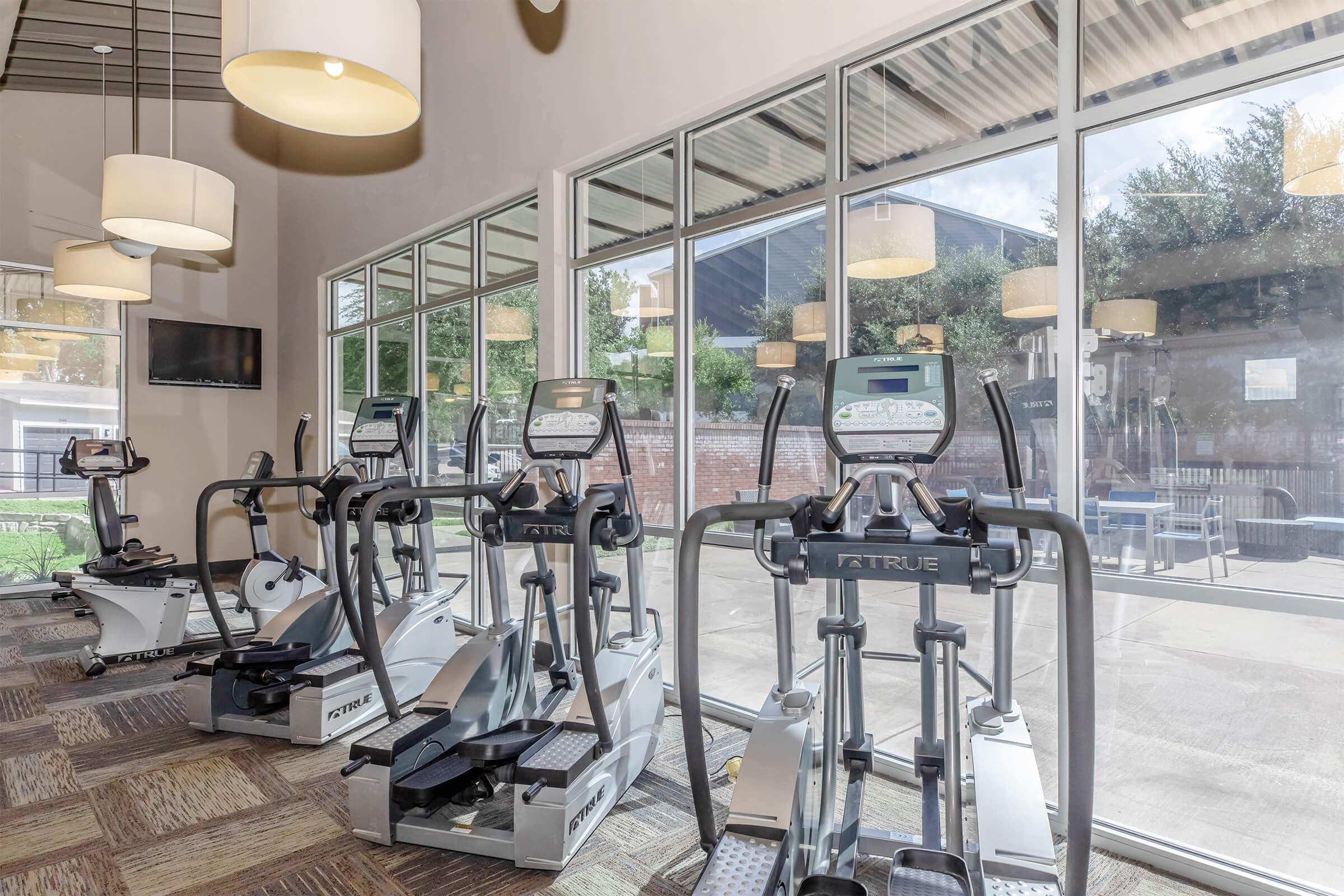 A bright and modern gym space featuring several elliptical machines arranged in front of large windows that provide natural light. The flooring has a patterned design, and there are pendant lights hanging from the ceiling, contributing to a welcoming atmosphere.