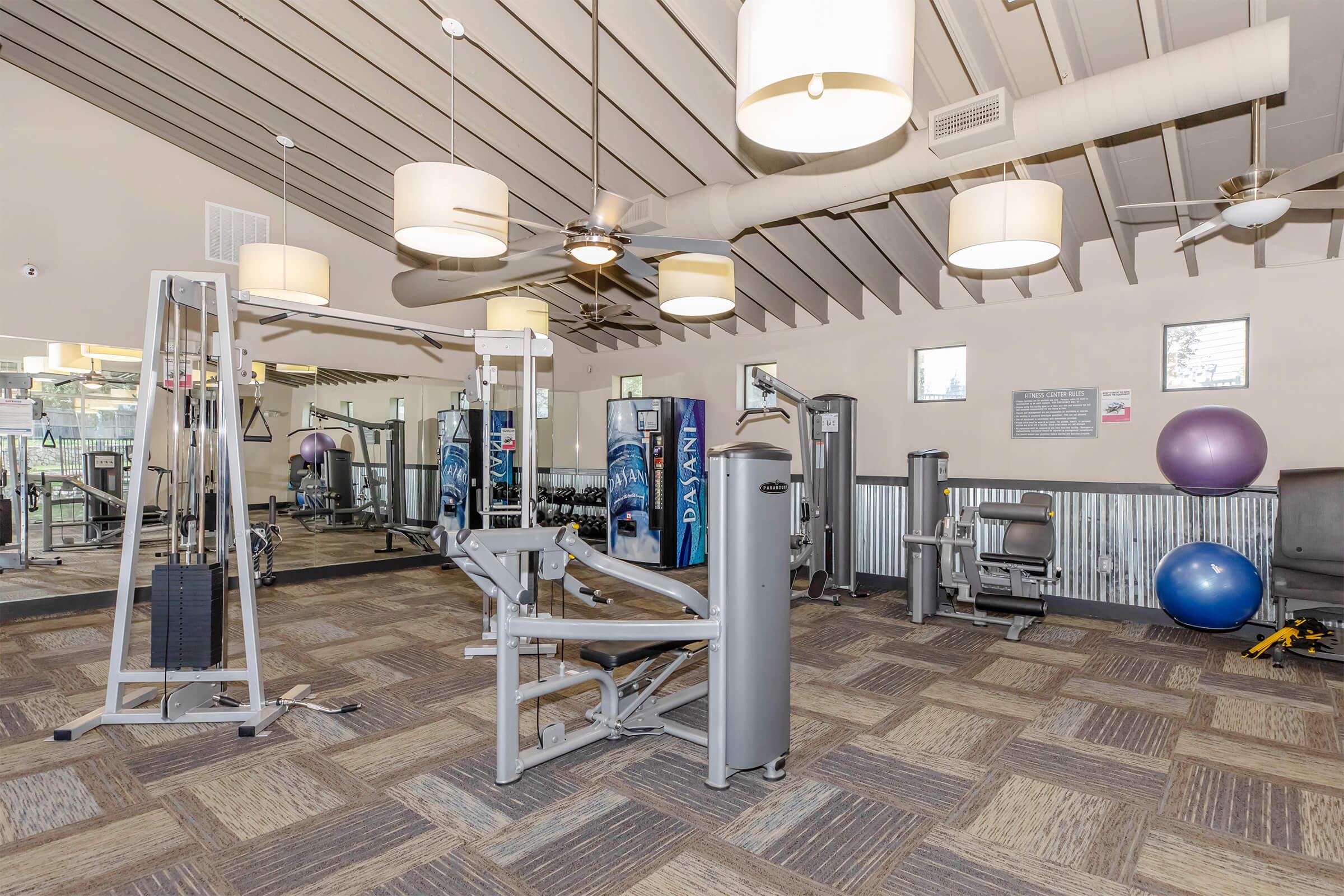 A spacious gym featuring various exercise equipment, including strength training machines and weights. The room has modern lighting, ceiling fans, large mirrors, and fitness accessories like exercise balls, providing a bright and inviting workout environment.
