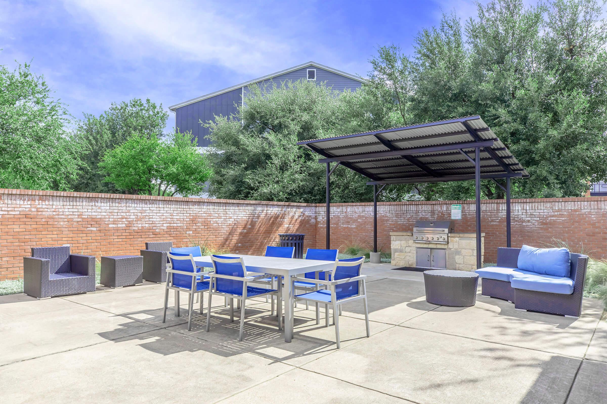 An outdoor patio area featuring a metal table with blue chairs, a lounge space with blue cushions, and a barbecue grill under a shaded pergola. Surrounding greenery and a red brick wall create a relaxing atmosphere.