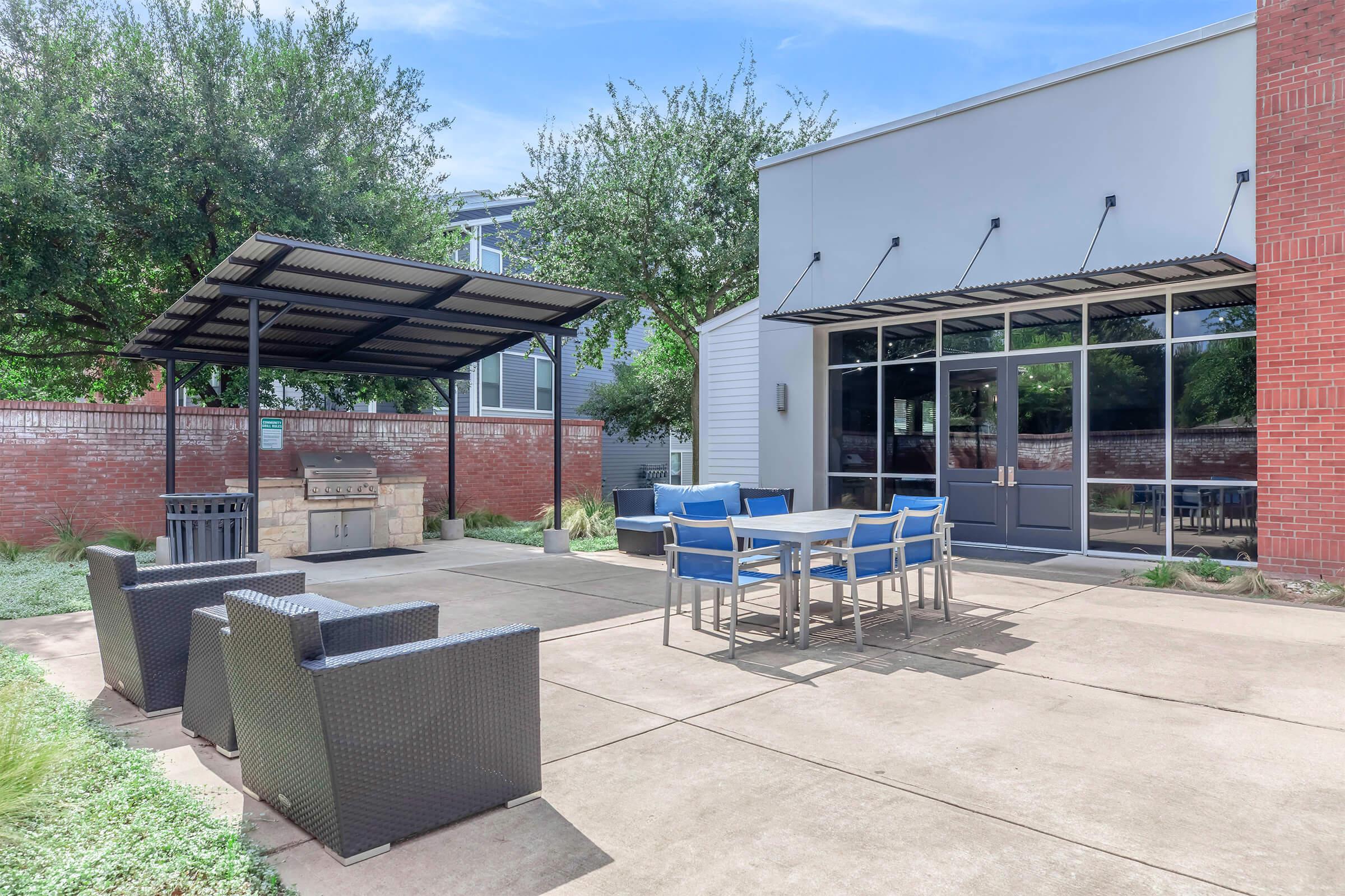 Outdoor patio area featuring a grilling station, a table with blue chairs, and wicker seating. Surrounded by greenery and trees, with a modern building nearby. Ideal for gatherings and relaxation. Clear blue sky enhances the inviting atmosphere.