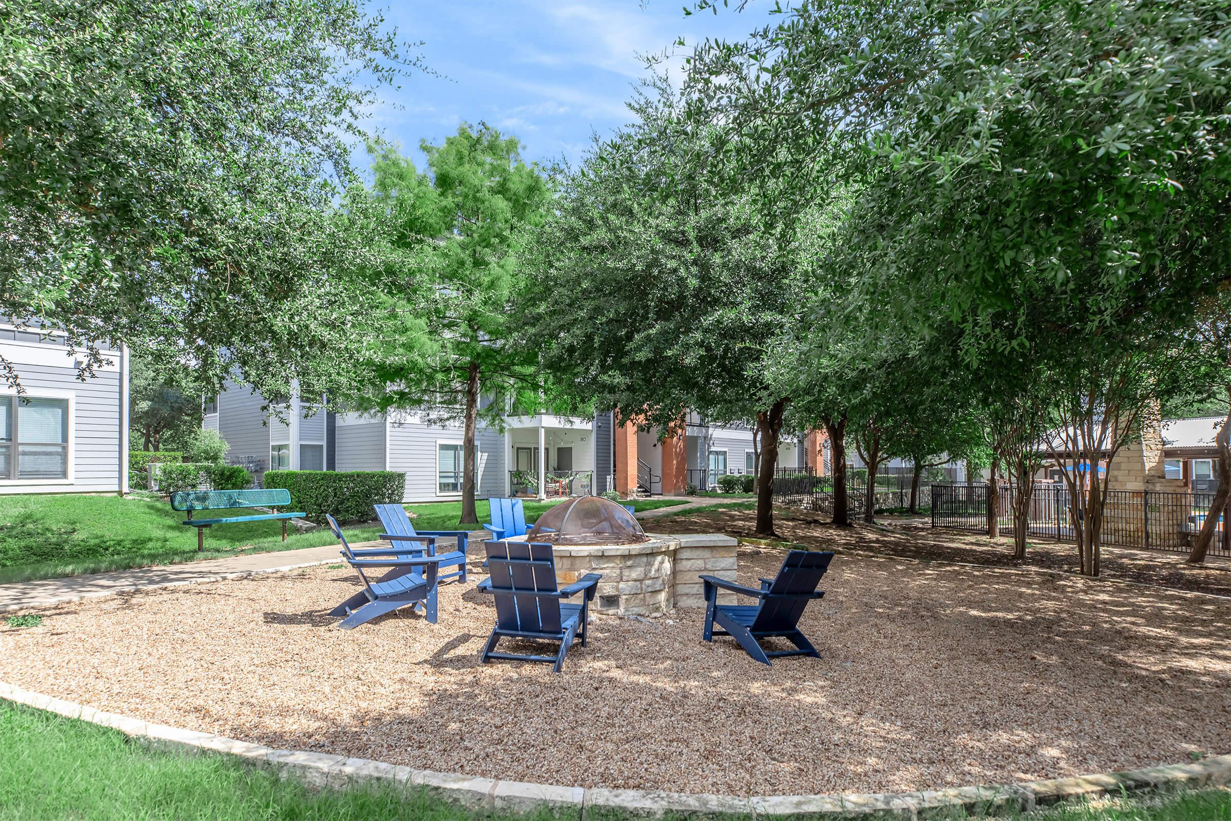 A serene outdoor recreational area featuring a circular stone fire pit surrounded by several blue chairs, set on a bed of gravel. Lush green trees provide shade, with nearby benches visible in a tranquil park-like setting. Ideal for gatherings and relaxation.
