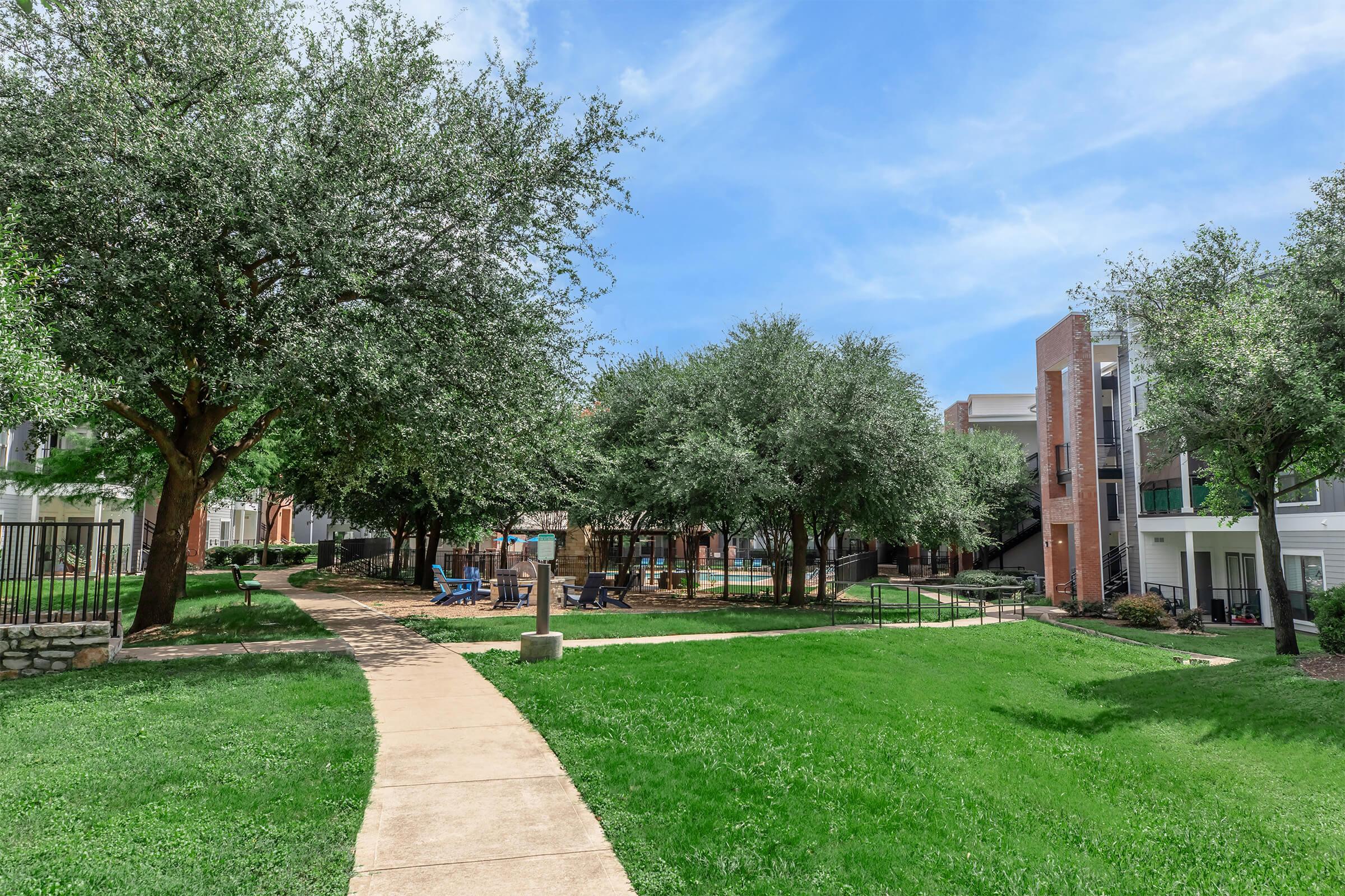 Lush green landscape featuring well-maintained lawns and trees surrounding a pathway. On one side, modern residential buildings are visible, and a small recreational area with seating is present. The sky is clear with a few clouds, creating a bright and inviting atmosphere.