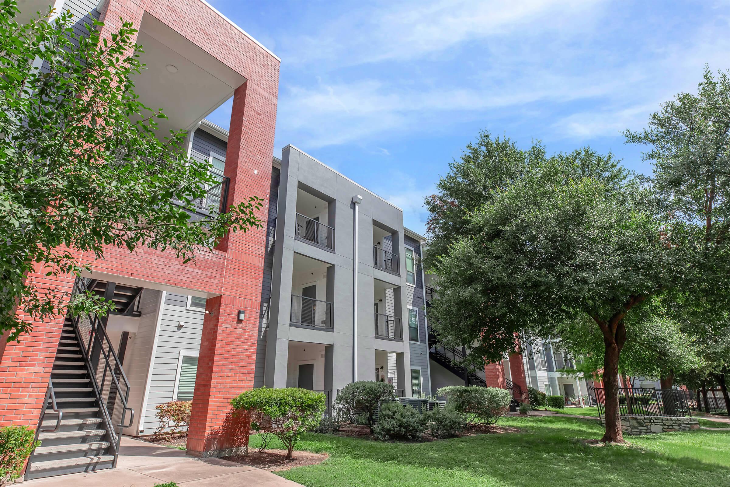 Apartment complex with modern architecture, featuring red brick and gray buildings. Lush green landscaping surrounds the buildings, including trees and shrubs. A staircase leads to the upper floors of one of the buildings, under a clear blue sky.