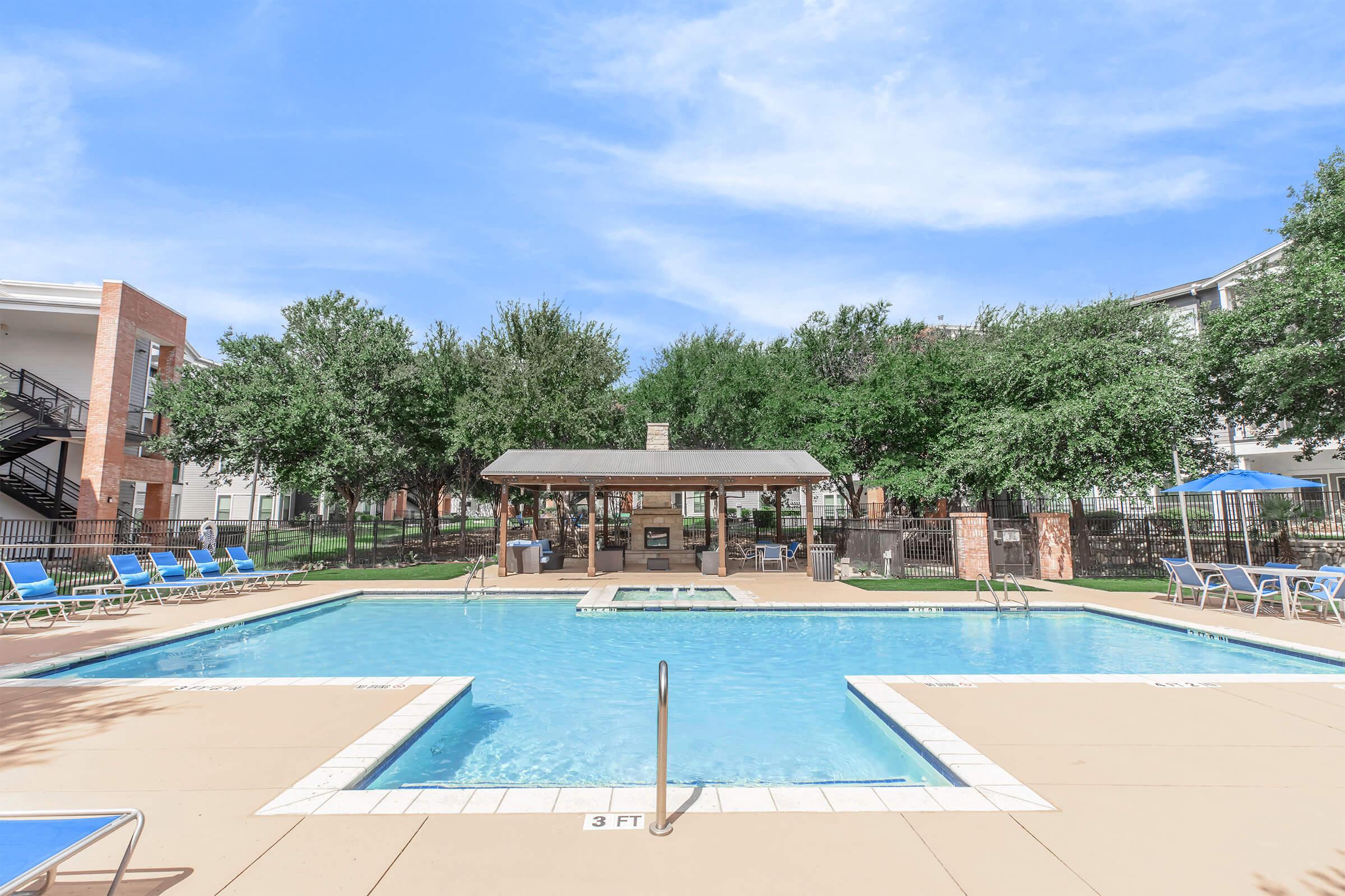 A clear swimming pool surrounded by lounge chairs and trees on a sunny day. There is a shaded gazebo area near the pool, and the surrounding buildings are visible in the background. The pool features a shallow end marked with "3 FT."