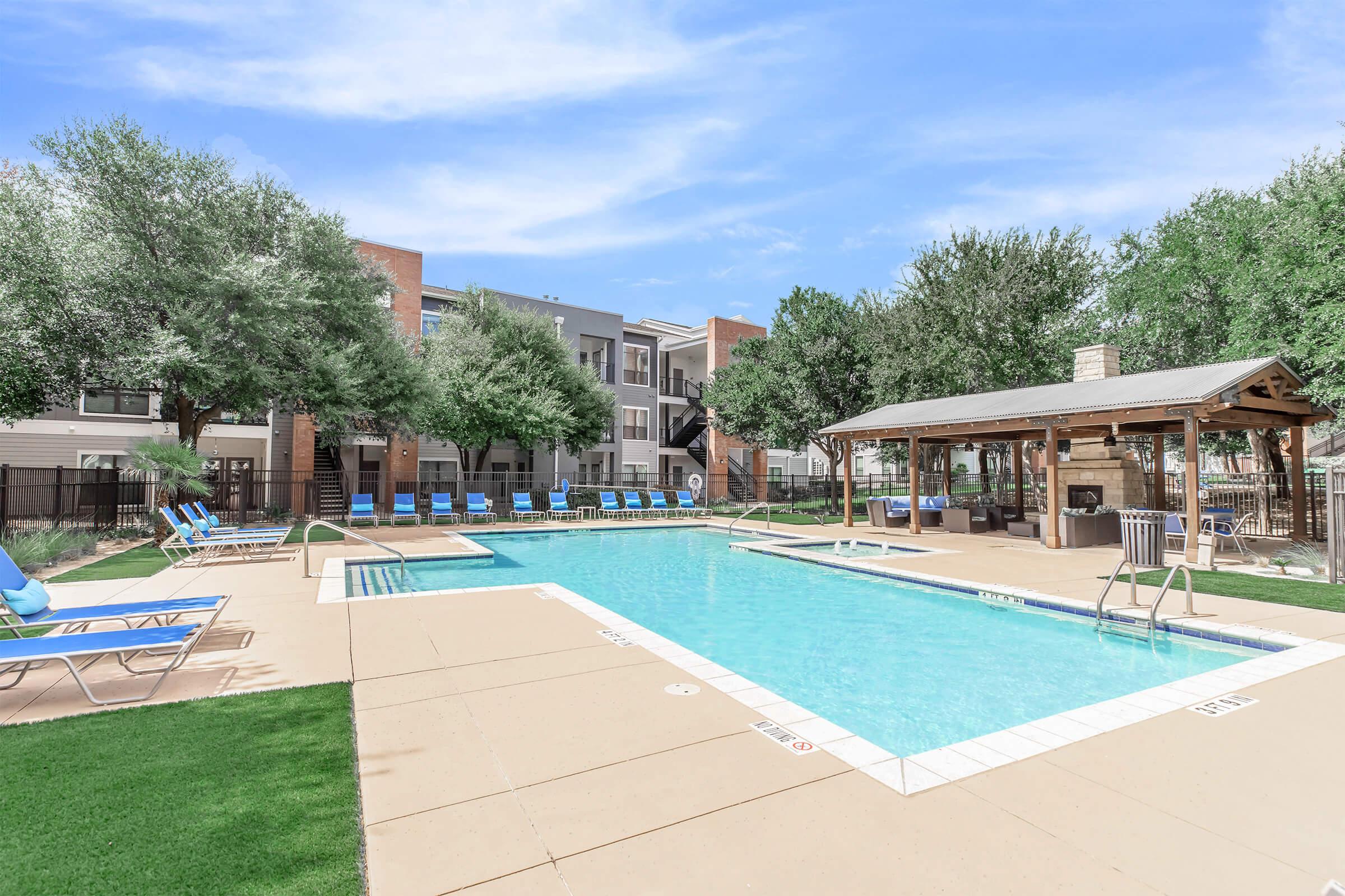 A sunny outdoor swimming pool surrounded by lounge chairs and shaded by trees. There is a gazebo nearby with picnic tables and a barbecue grill, all set in a well-maintained apartment complex. The sky is clear and blue, enhancing the inviting atmosphere.