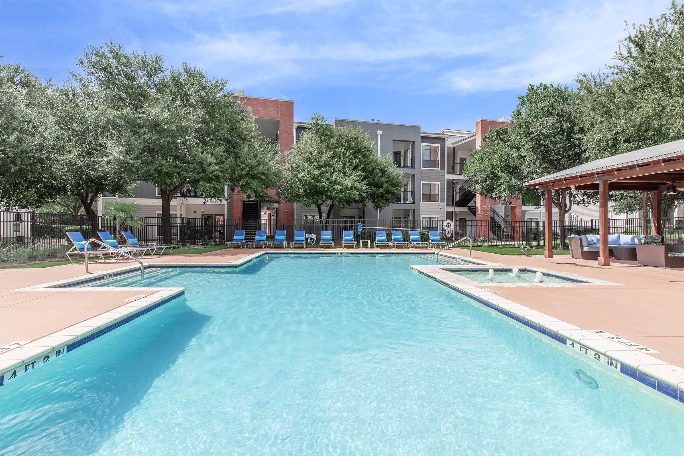 A clear swimming pool with lounge chairs around it, surrounded by green trees and modern apartment buildings in the background. The sky is bright with a few clouds, creating a pleasant outdoor atmosphere. A shaded area with a gazebo is visible near the poolside.