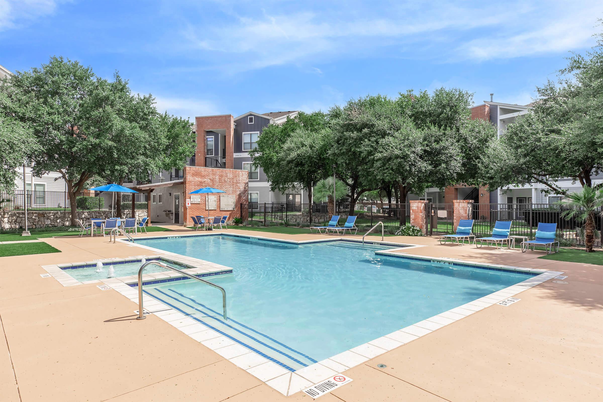 A swimming pool surrounded by lounging chairs and shaded by trees, with umbrellas positioned nearby. The area is part of an apartment complex, featuring well-maintained landscaping and a clear blue sky above. The pool has steps leading in and a clean, inviting appearance.