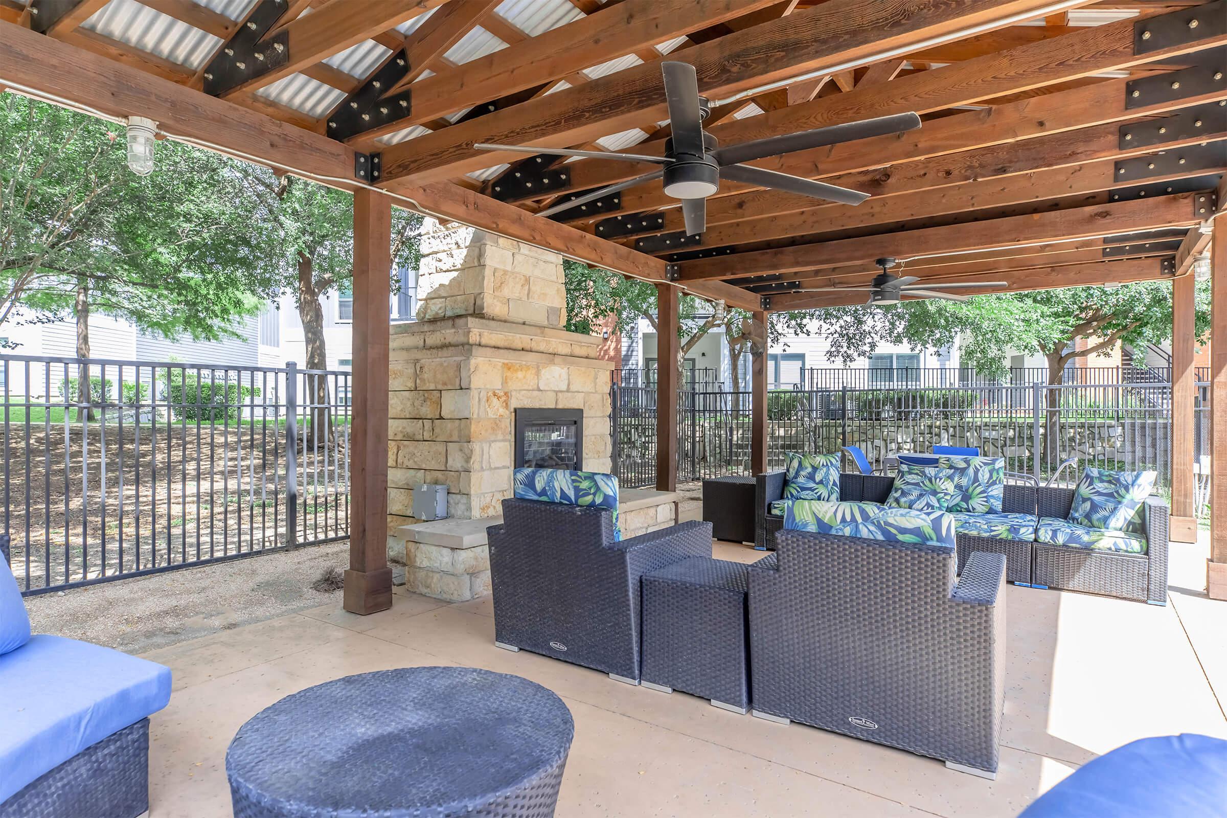 Outdoor seating area with a covered pergola, featuring wicker furniture with tropical-patterned cushions, a stone fireplace, and ceiling fans. The space is surrounded by greenery and has a fenced area for added privacy.