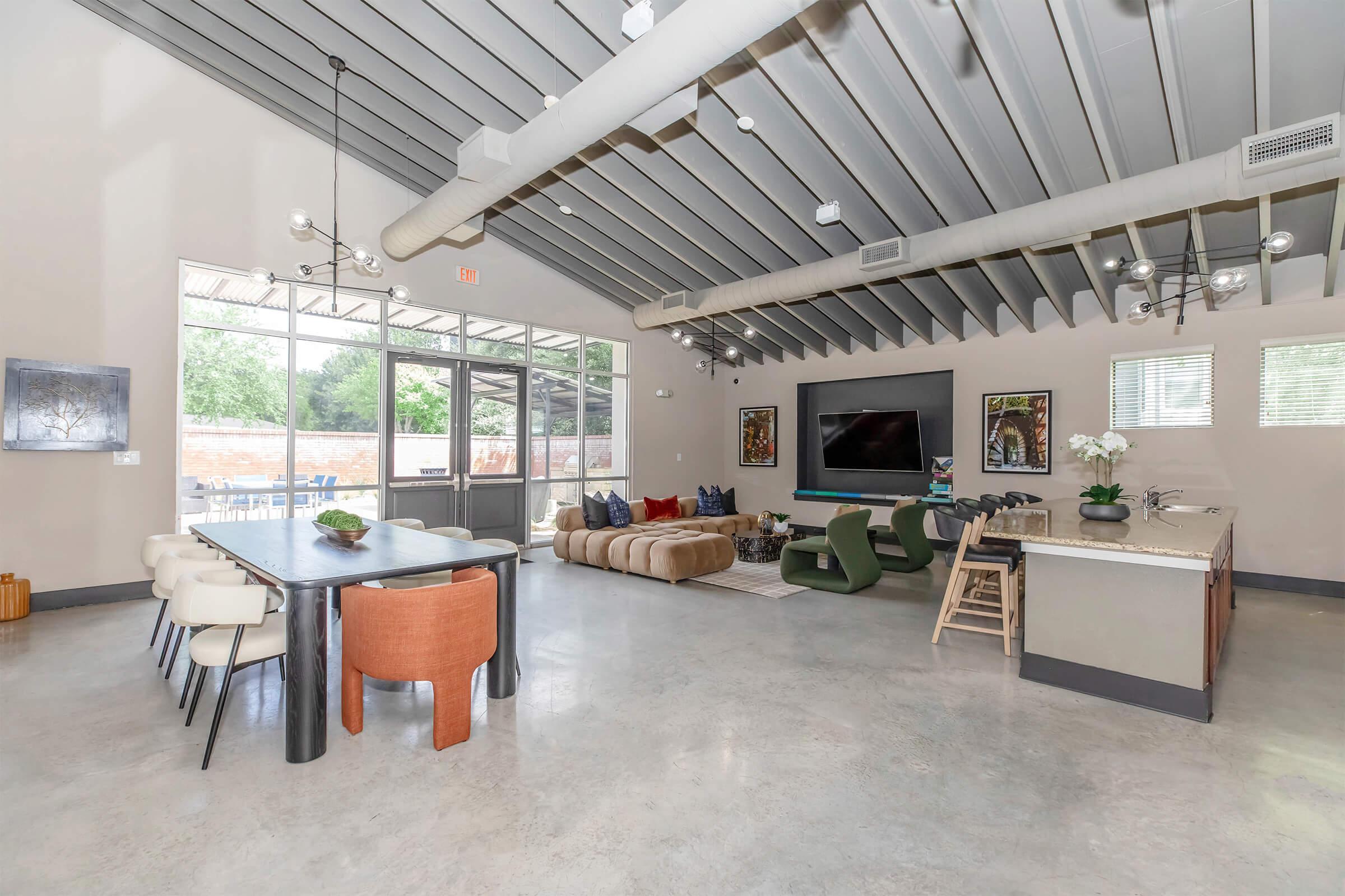 Bright, modern living space featuring a spacious layout with high ceilings, large windows, and concrete flooring. It includes a dining area with a long table and colorful chairs, a cozy seating area with a sectional couch, and a kitchen with a bar. Natural light fills the room, enhancing the contemporary design.