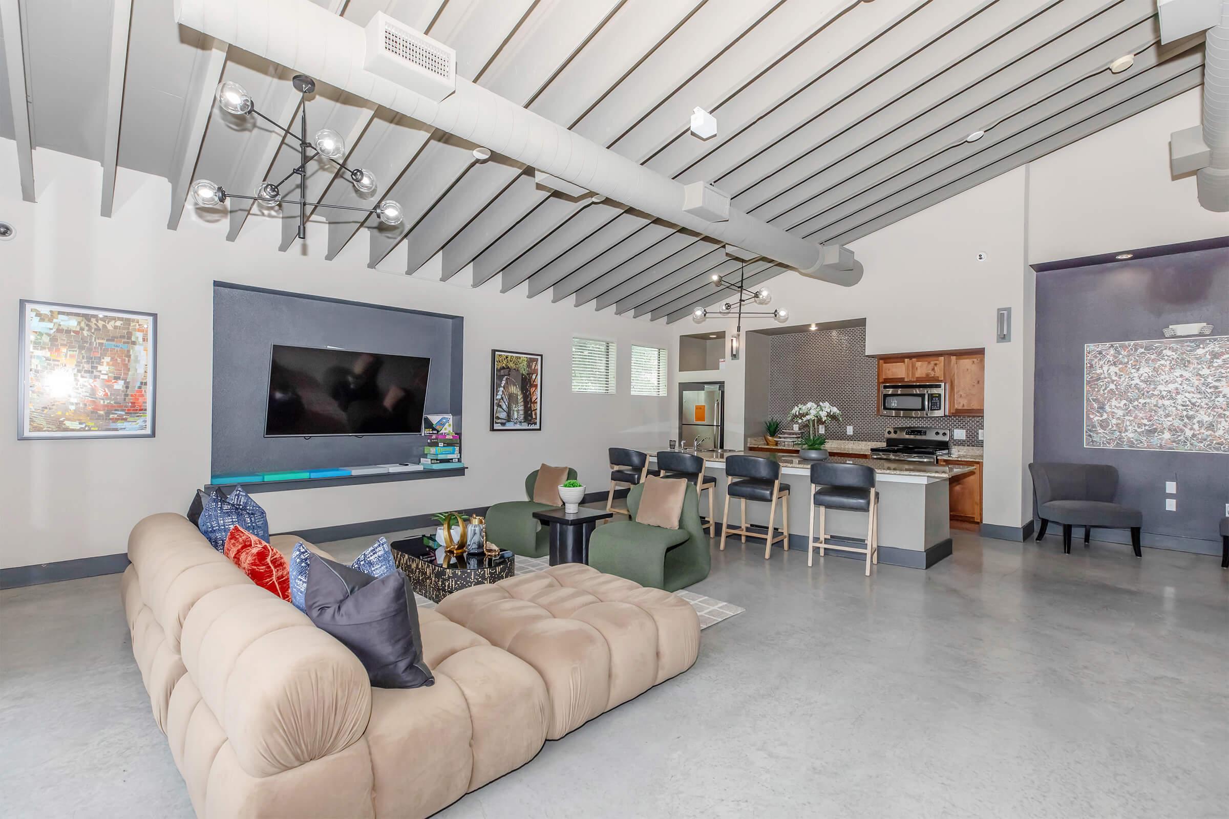 Spacious modern living room featuring a beige sectional sofa with colorful cushions, a large flat-screen TV mounted on the wall, and a stylish coffee table. A dining area with bar stools is visible, along with contemporary decor and pendant lighting, showcasing a clean, open layout with gray walls and a polished concrete floor.