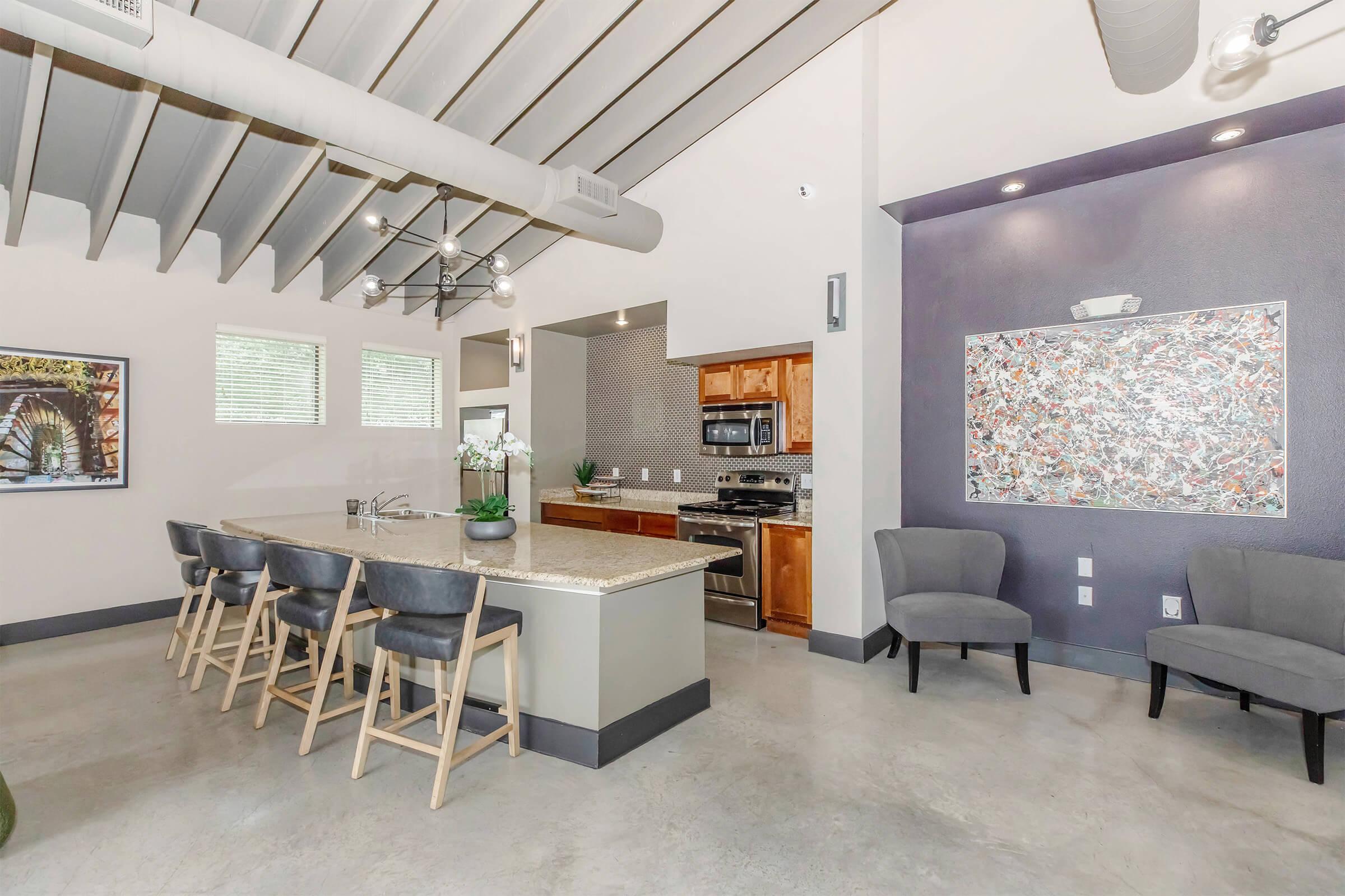 Modern kitchen interior featuring a large island with barstools, stainless steel appliances, and sleek cabinetry. The space has high ceilings with exposed beams, windows for natural light, and a decorative wall art piece. The flooring is polished concrete, creating a contemporary feel.