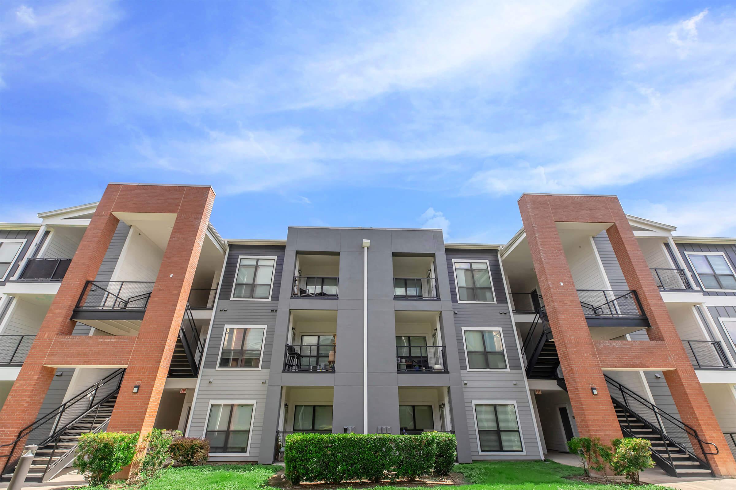 A modern apartment building featuring two prominent brick pillars, multiple balconies, and a well-maintained landscape. The sky is bright and clear, enhancing the contemporary aesthetic of the architecture.
