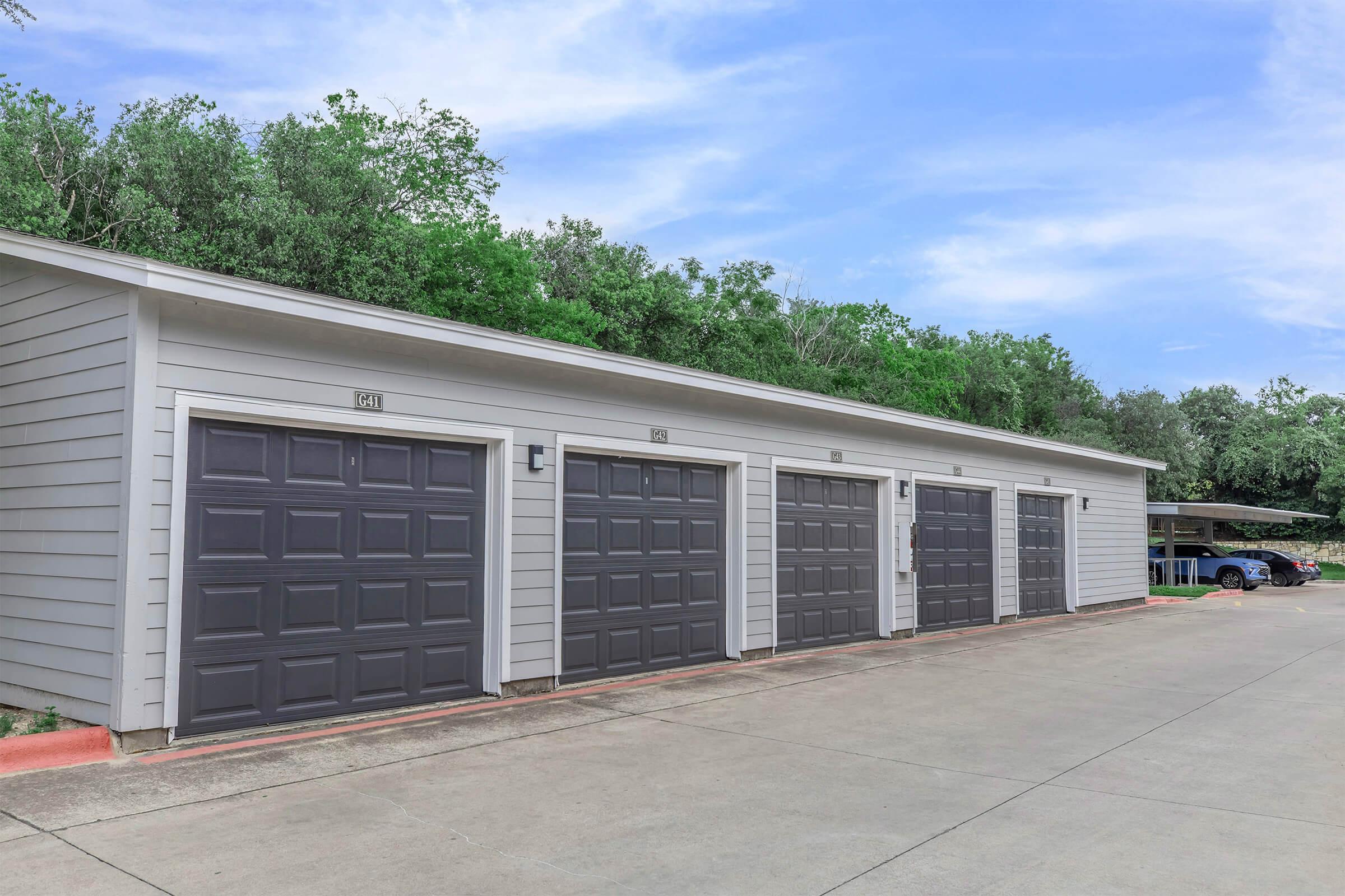 A row of three garage doors with a light gray exterior, surrounded by greenery. The garages are part of a single-story building with a paved driveway, and a vehicle is parked nearby in the shaded area. The sky above is clear with a few clouds.