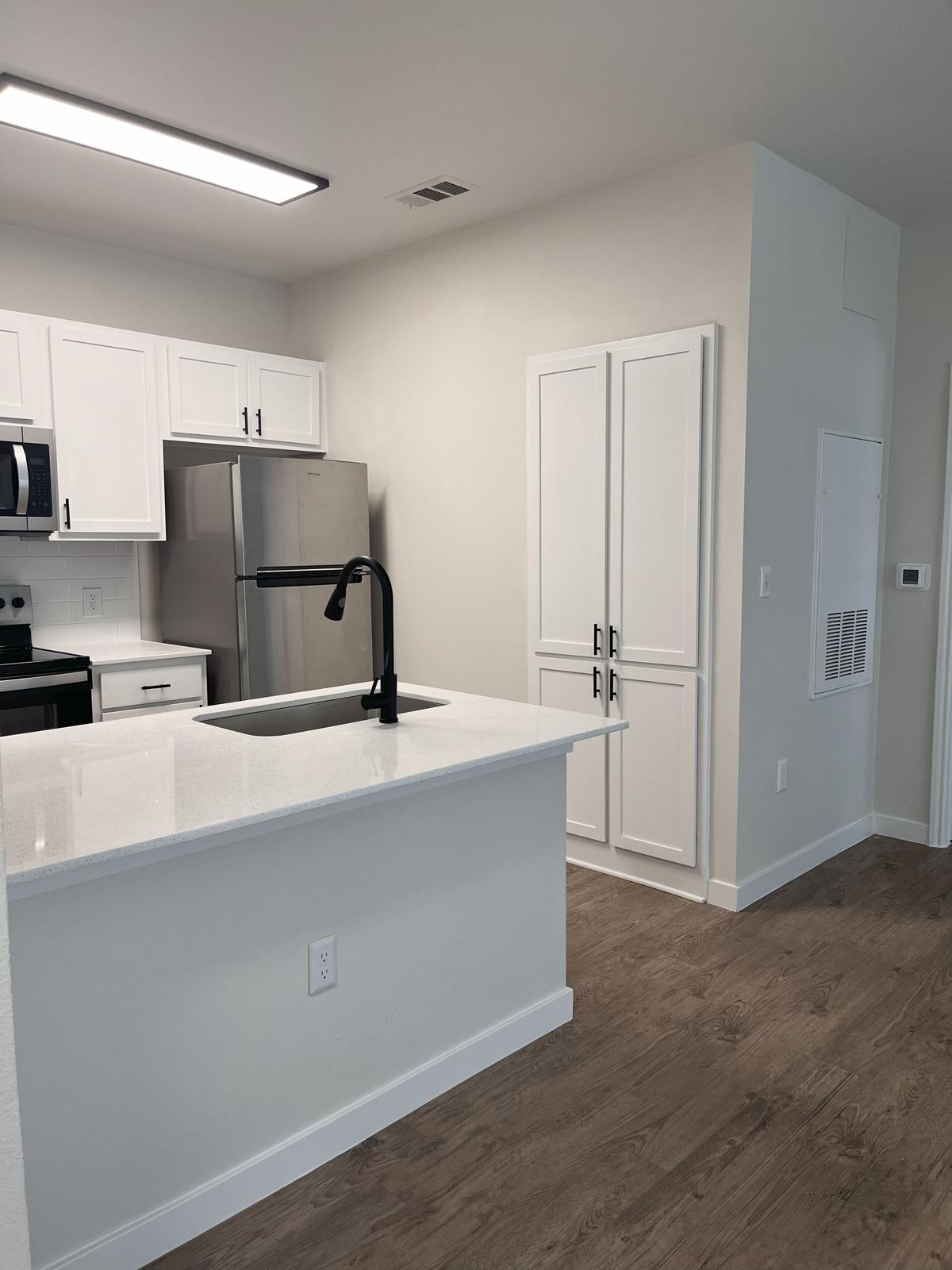 Modern kitchen interior featuring white cabinets, a stainless steel refrigerator, and a sleek black faucet above a white countertop. The open layout includes a wooden floor and a well-lit space, with appliances and storage units visible. Natural light brightens the area, creating an inviting atmosphere.