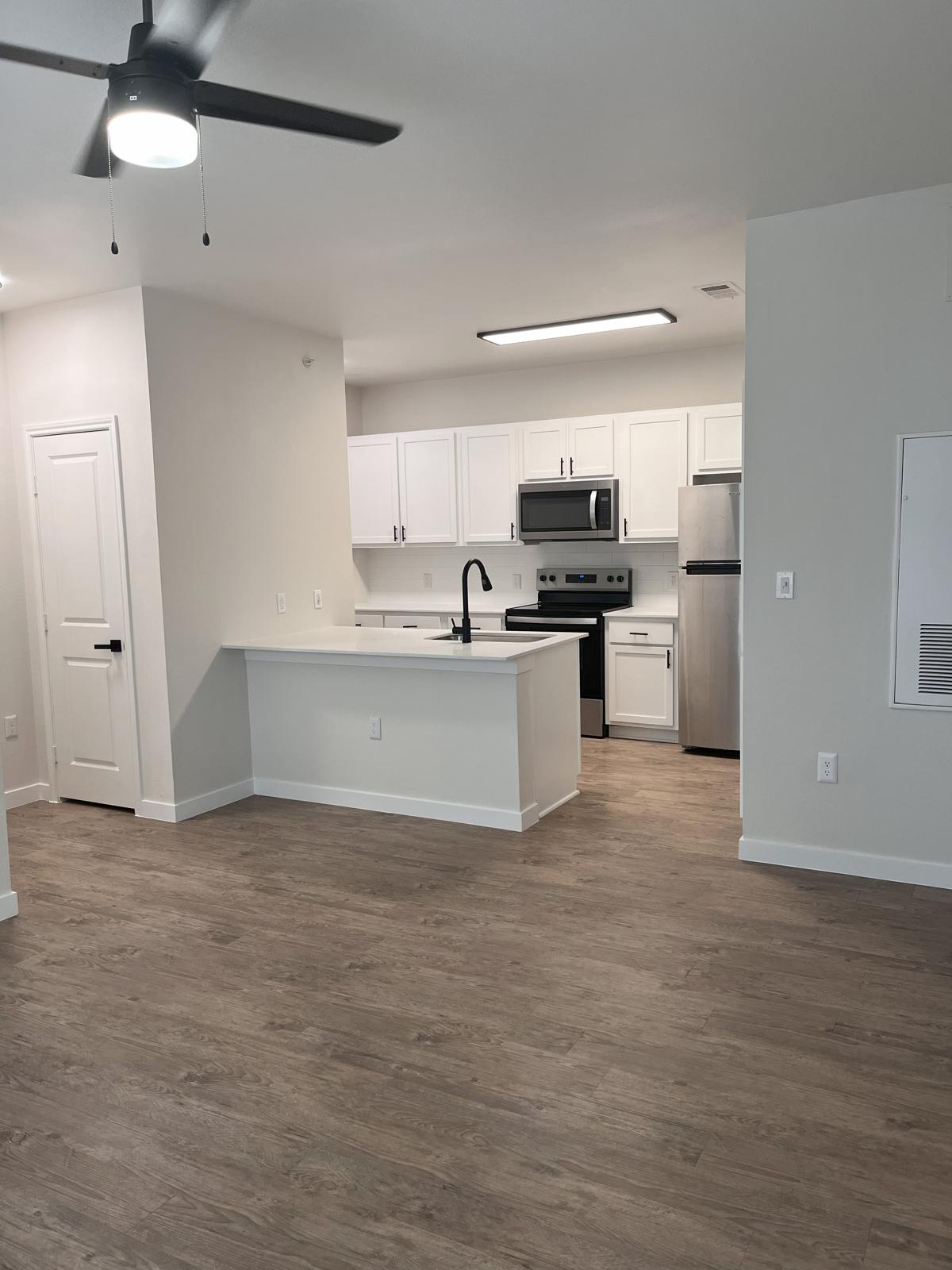 A modern, open-concept kitchen and living area featuring white cabinetry, stainless steel appliances, and a light wood-style floor. The kitchen island includes a sink and is adjacent to the living space, which has a ceiling fan and is well-lit with natural light. The walls are painted in a neutral color.