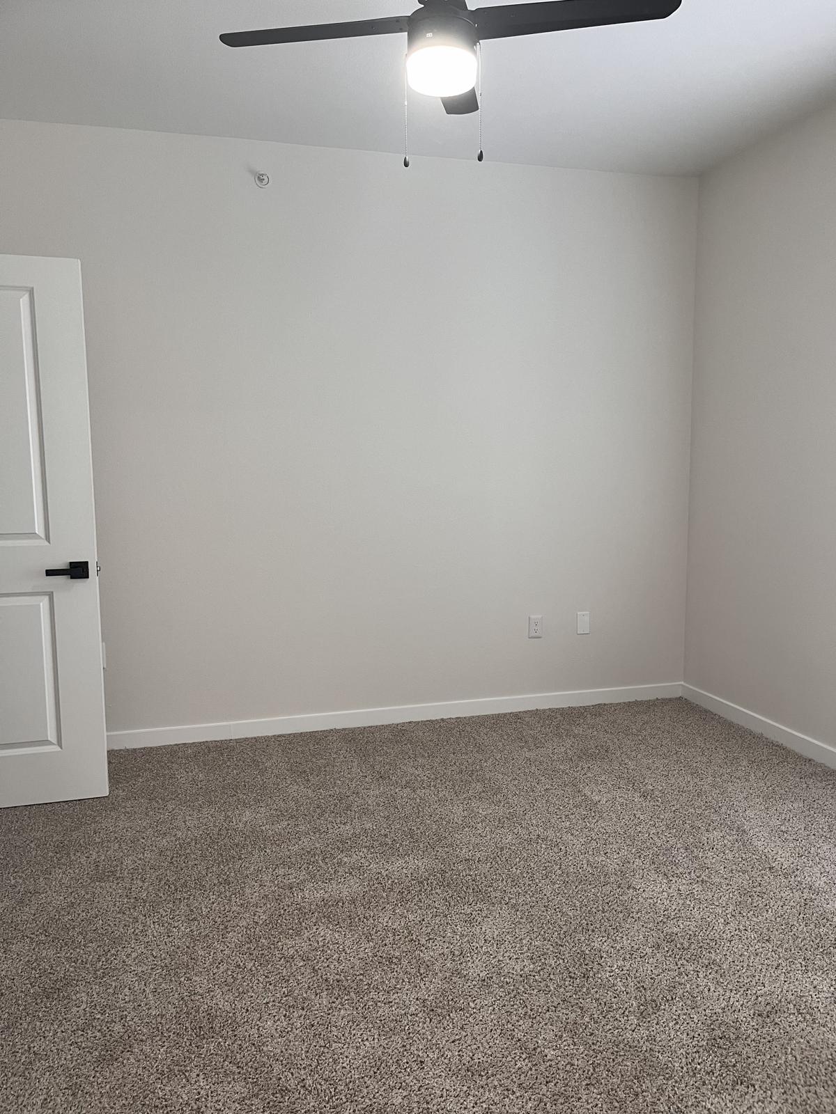A clean, empty room with light beige carpet and light gray walls. A ceiling fan is mounted in the center, and there is a closed white door on the left side. Natural light enhances the spacious feel of the area, making it suitable for various uses.