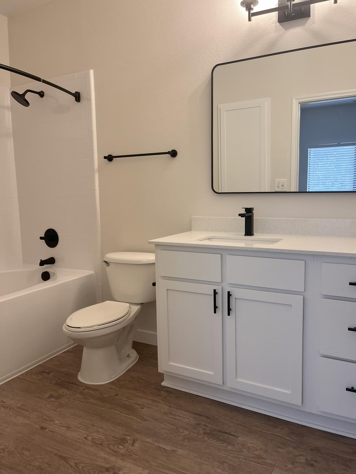 A modern bathroom featuring a white bathtub with black fixtures, a toilet, a white vanity with a sink, and a large mirror. The walls are light-colored, and there is a dark towel rack and light fixture. A window provides natural light in the background. The flooring is a warm wood finish.