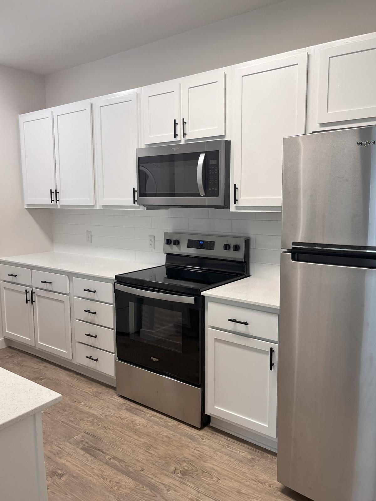 A modern kitchen featuring white cabinets with black handles, a stainless steel refrigerator, a stainless steel microwave above a black electric range, and a white countertop. The flooring is light wood, and the walls are painted in a light color, creating a clean and contemporary look.