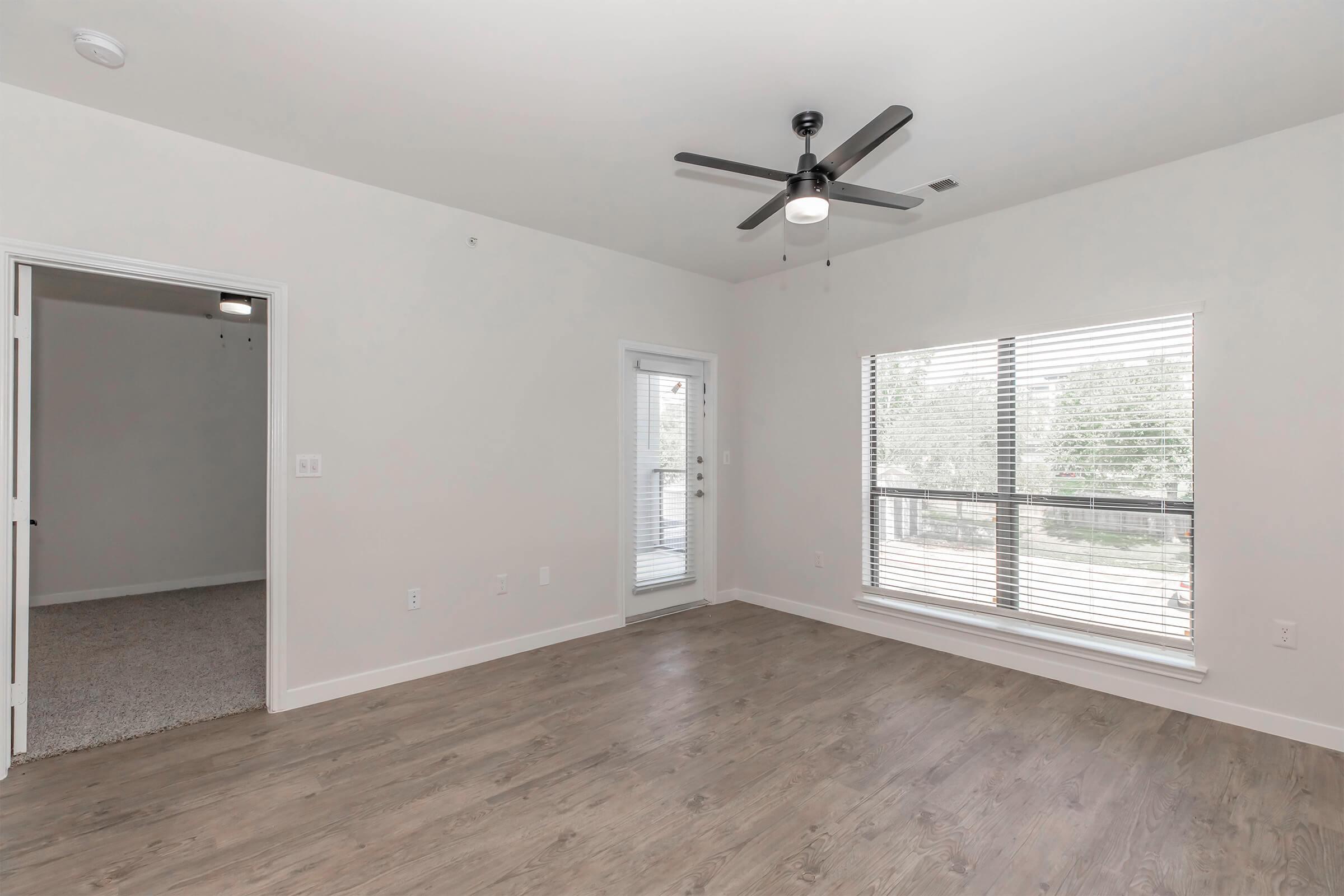 A spacious, modern room with light wood flooring, large windows featuring white blinds, and a ceiling fan. There is a door leading to a small balcony and an open doorway leading to another room. The walls are painted in a neutral color, creating a bright and airy atmosphere.