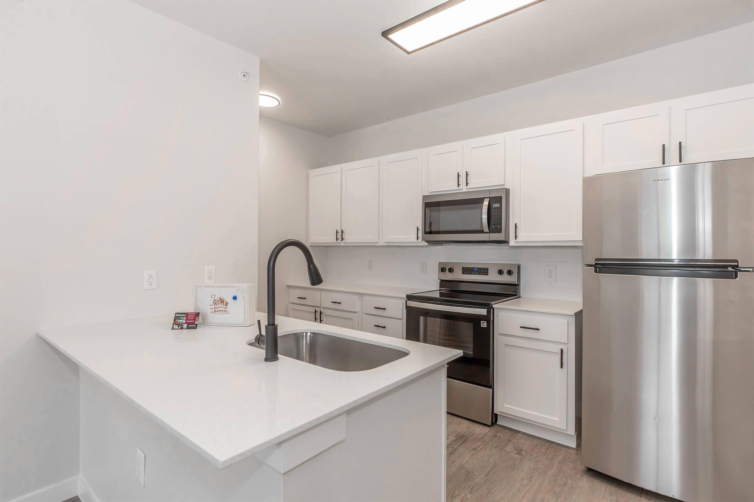 Modern kitchen featuring white cabinets, stainless steel appliances, a sleek refrigerator, and a microwave. The countertop is made of white material, with a dark faucet above a spacious sink. Natural light illuminates the room, highlighting the minimalist design and clean lines.
