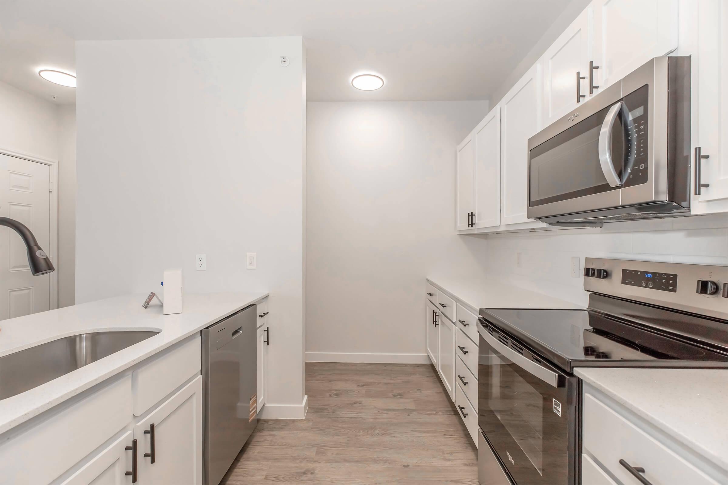 A modern kitchen featuring white cabinetry, stainless steel appliances including a microwave, oven, and dishwasher, along with a double sink. The space has light-colored laminate flooring and bright lighting, creating a clean and airy atmosphere.