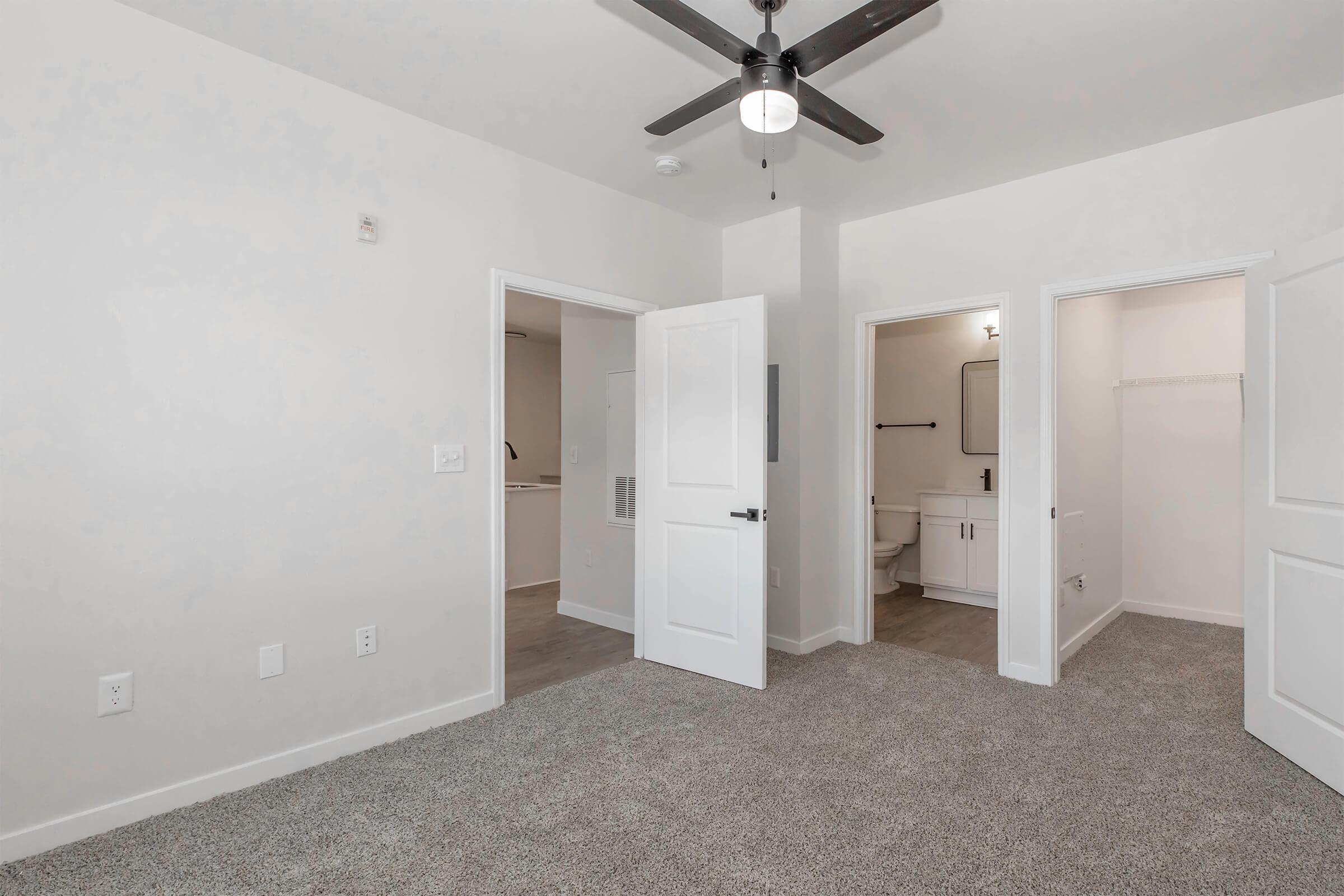 A well-lit, empty room featuring light gray carpet and white walls. Two open doors lead to a bathroom and a closet. A ceiling fan is installed, and the space has a modern design with minimal furnishings, creating a clean and inviting atmosphere.