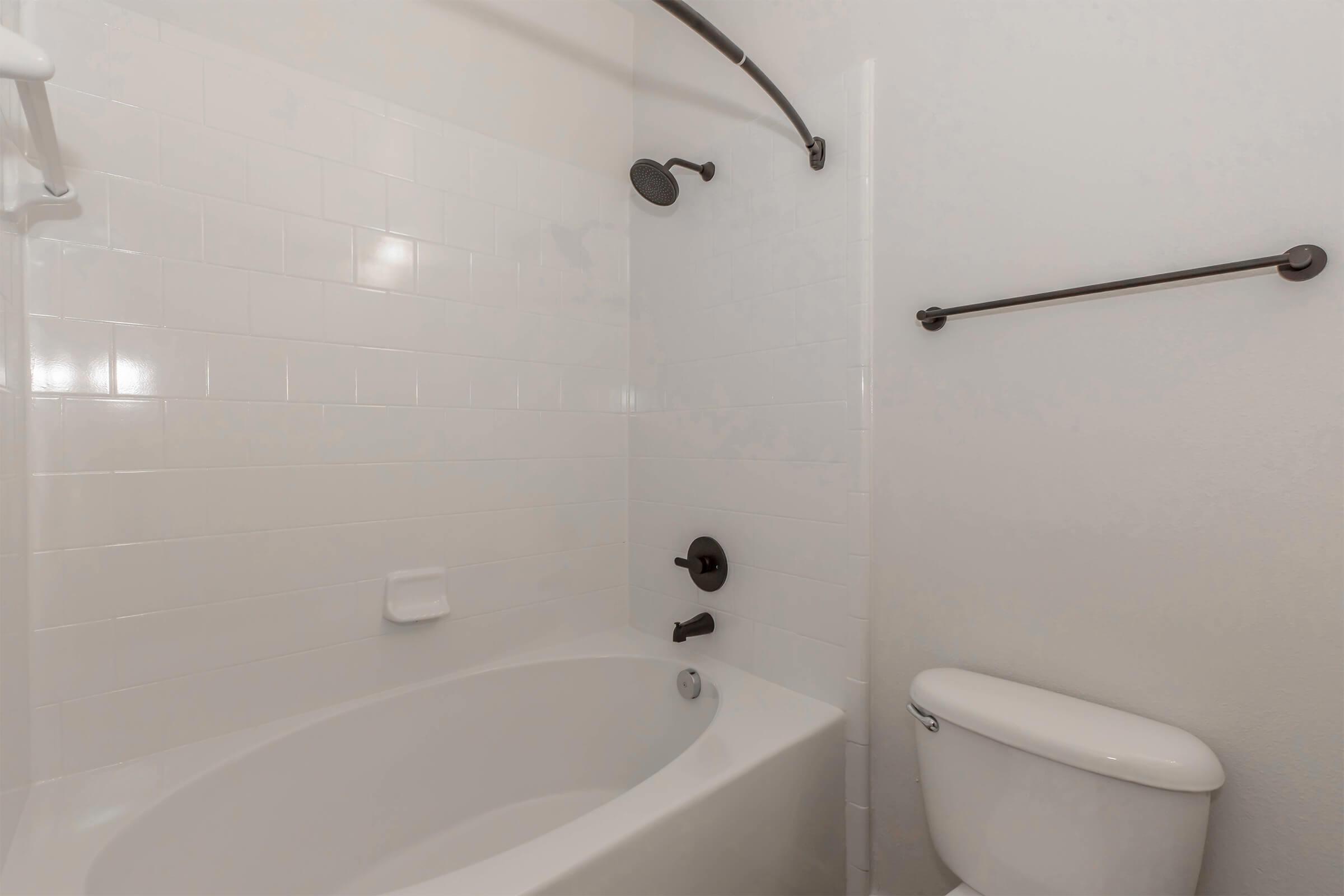 A clean, minimalist bathroom featuring a white bathtub with a showerhead above, white tile walls, and a brown towel bar. A toilet is positioned beside the bathtub, completing the simple, modern aesthetic of the space.