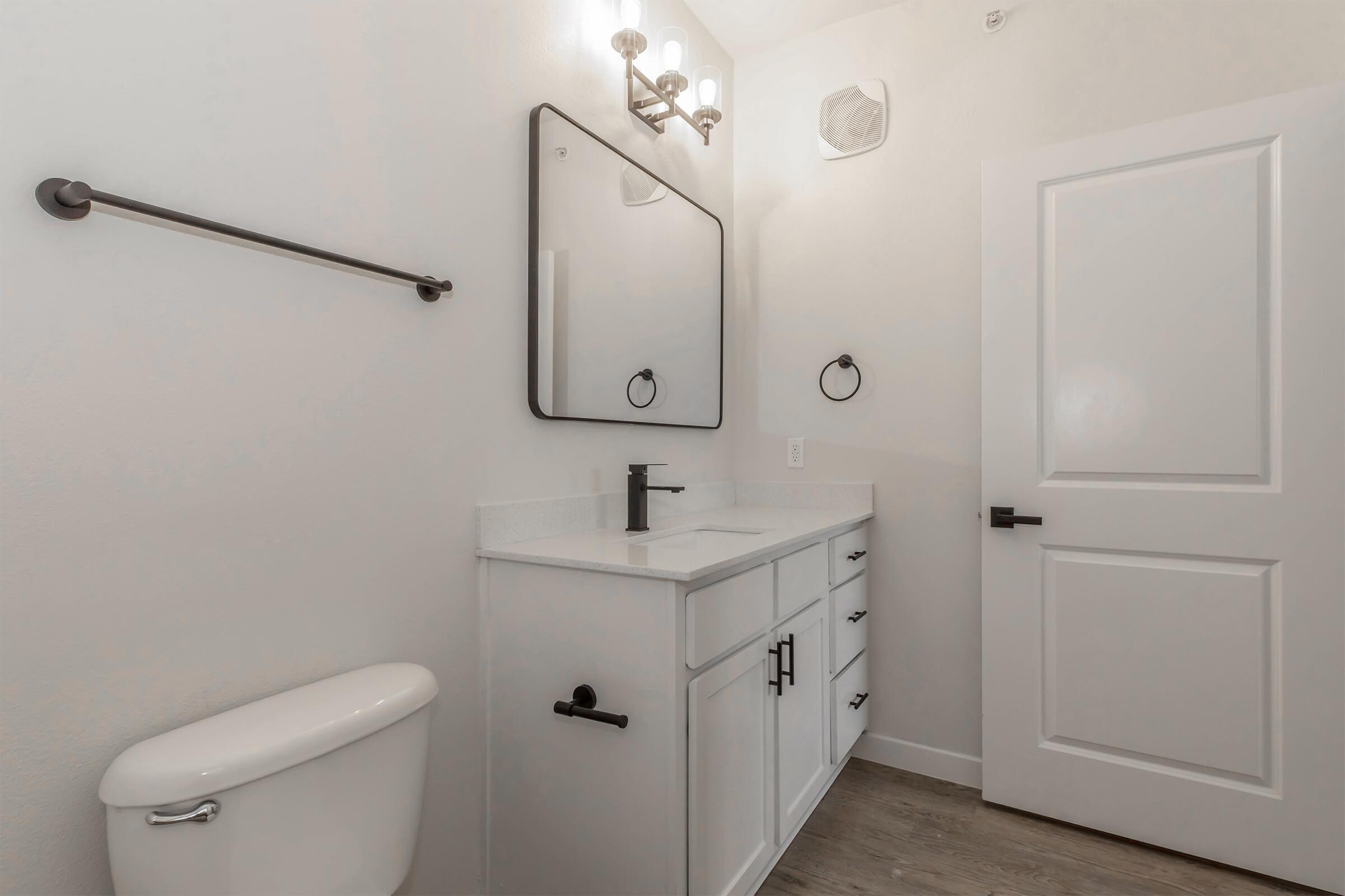 A modern bathroom featuring a white vanity with a sleek black faucet, a large wall mirror, a black towel rack, and a minimalist door. The walls are painted light, and there is a toilet in the corner along with a small vent. The flooring is a light wood look.
