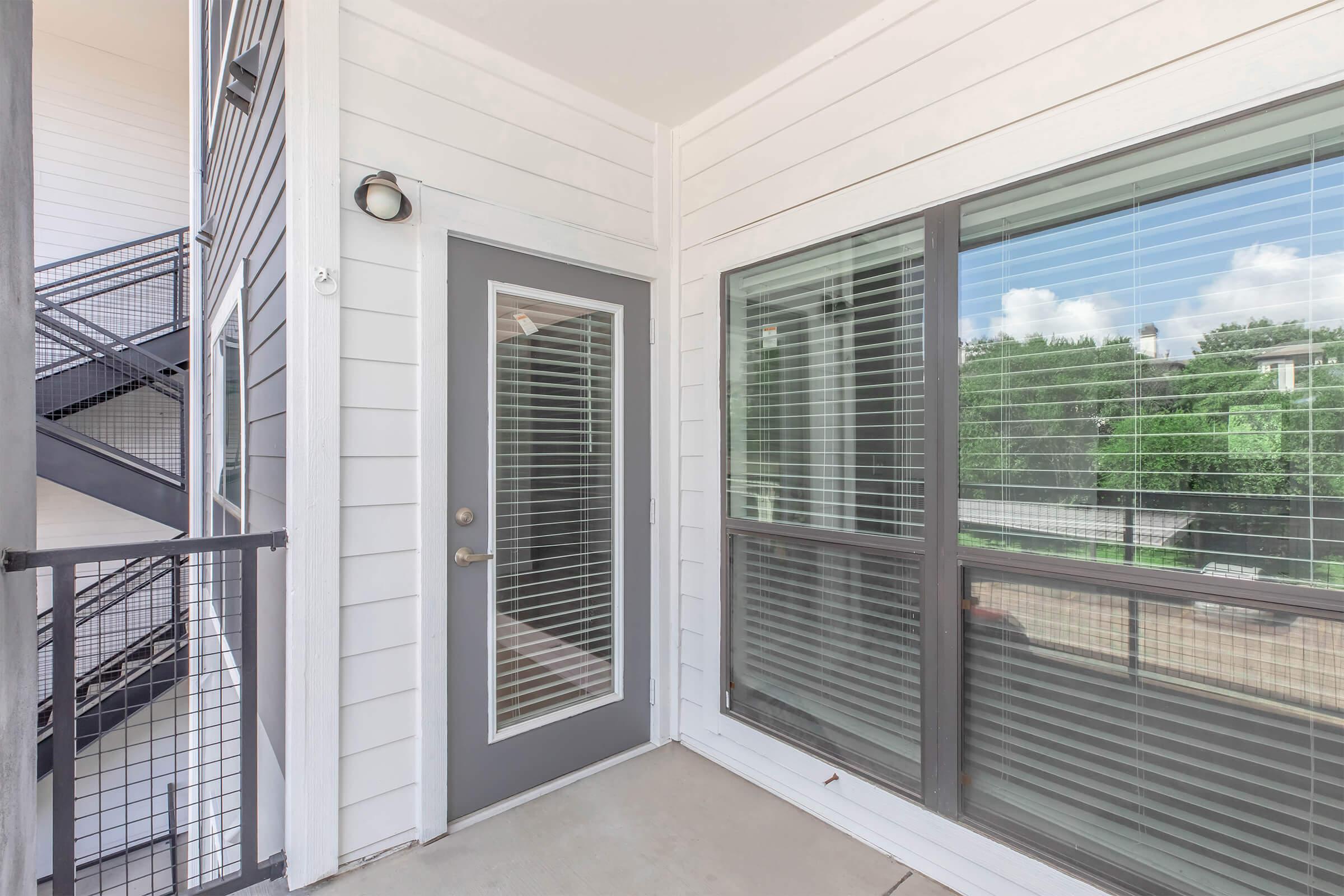 A view of an apartment entrance featuring a gray door, large windows with blinds, and a staircase visible in the background. The balcony area is well-lit with natural light, and the surrounding environment includes greenery.