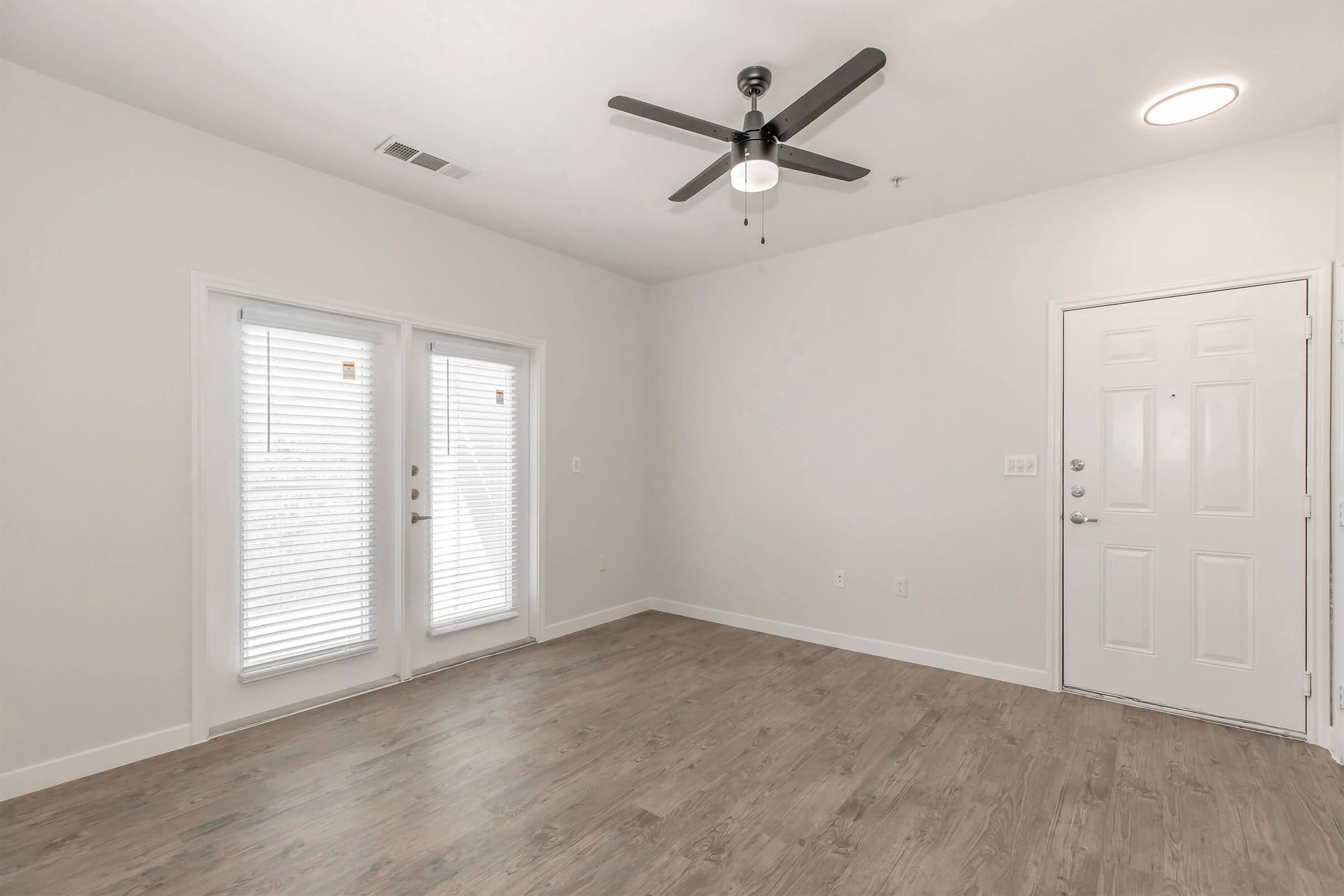 A spacious, empty room with light-colored walls and a ceiling fan. There are two large windows with blinds on one side, and a door leading outside. The flooring is a light wood laminate. The room has a clean and modern aesthetic, with a white door on the right side.