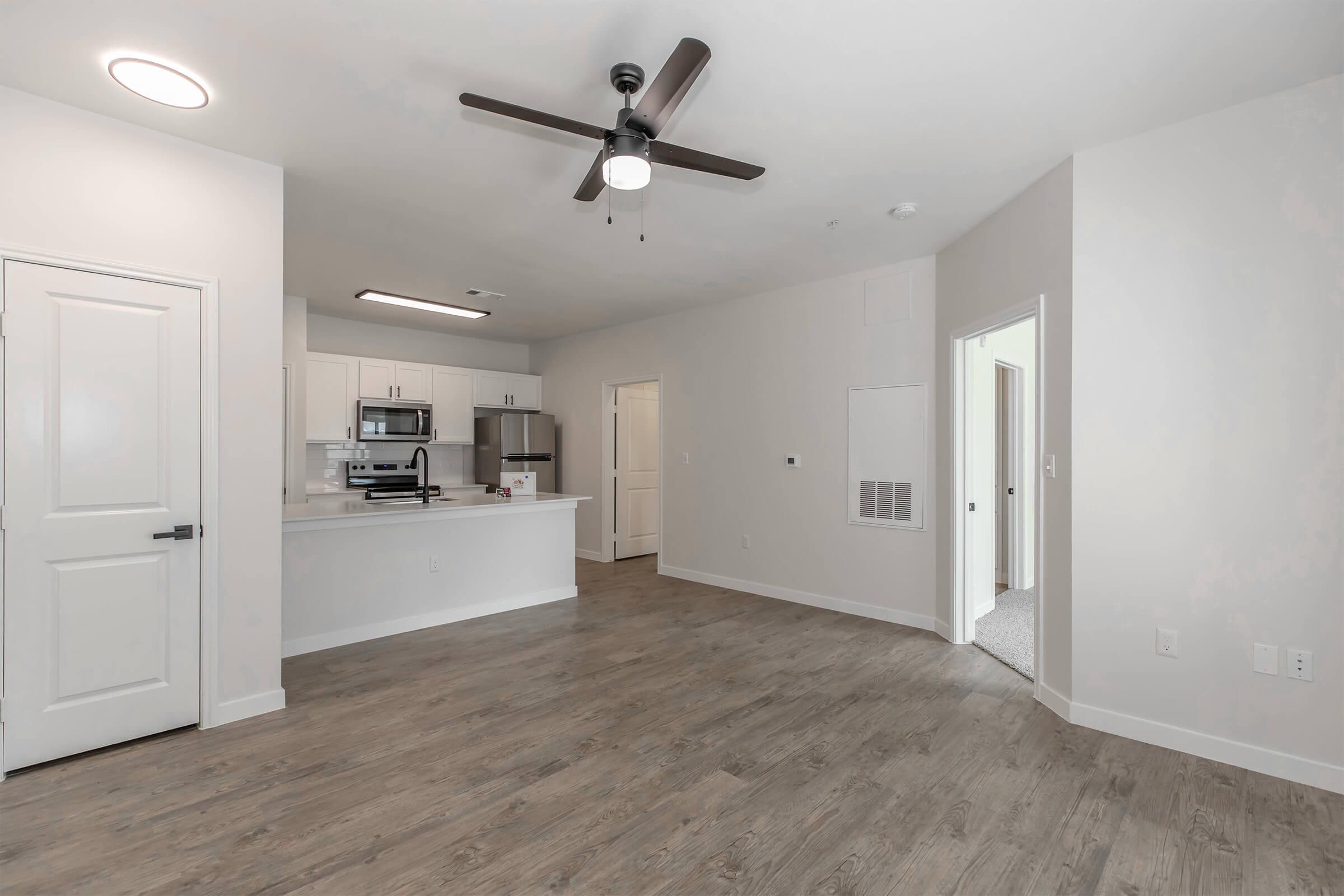 A modern living space featuring hardwood flooring, a ceiling fan, and an open layout. The kitchen area includes white cabinets and stainless steel appliances. There's a doorway leading to another room, and natural light illuminates the space, creating a warm and inviting atmosphere.