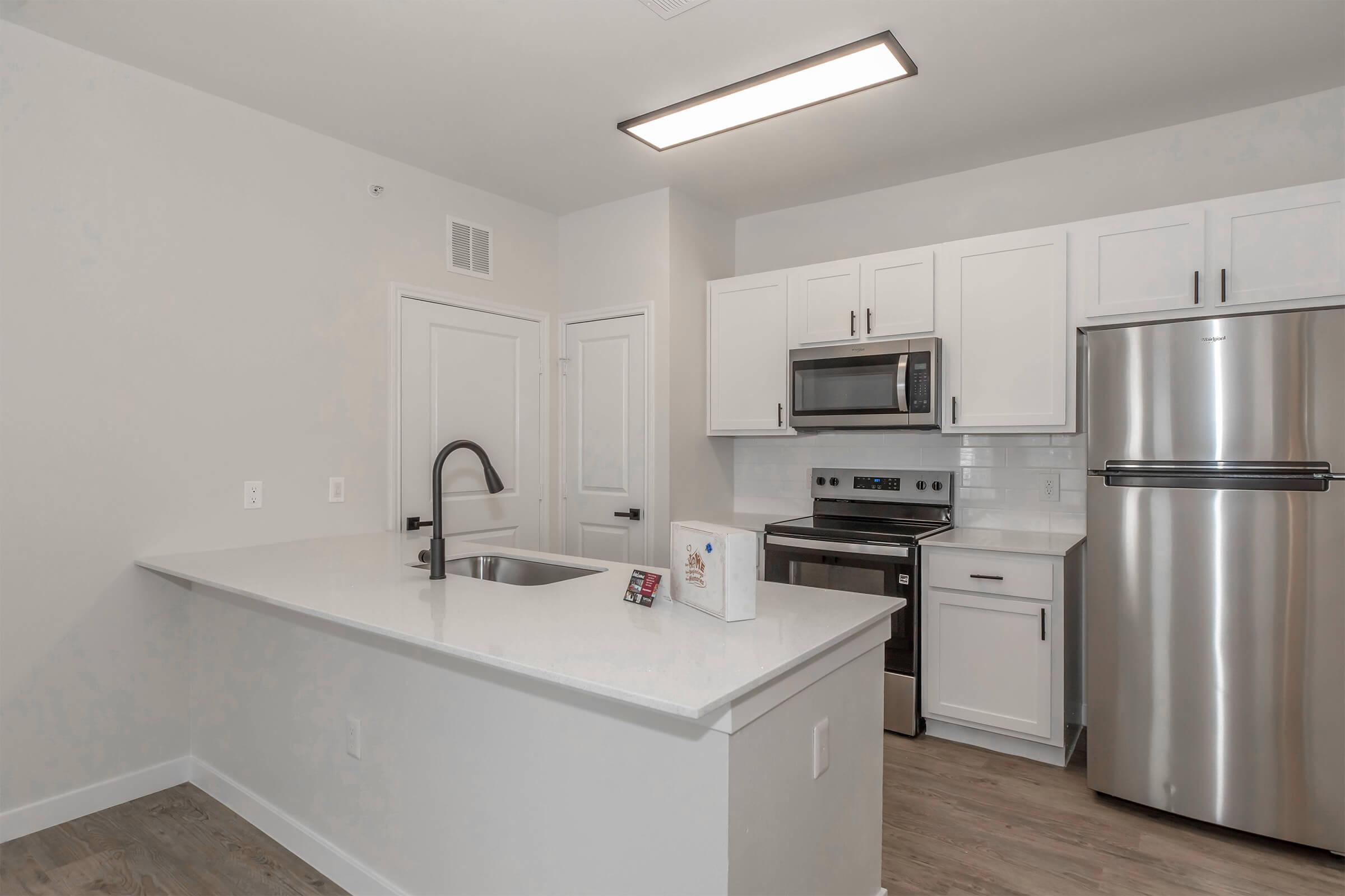 Modern kitchen featuring white cabinets, stainless steel appliances, and a large island with a sink. The countertop is light-colored, and there are ample cabinets for storage. Natural light streams in, highlighting the clean and contemporary design of the space.