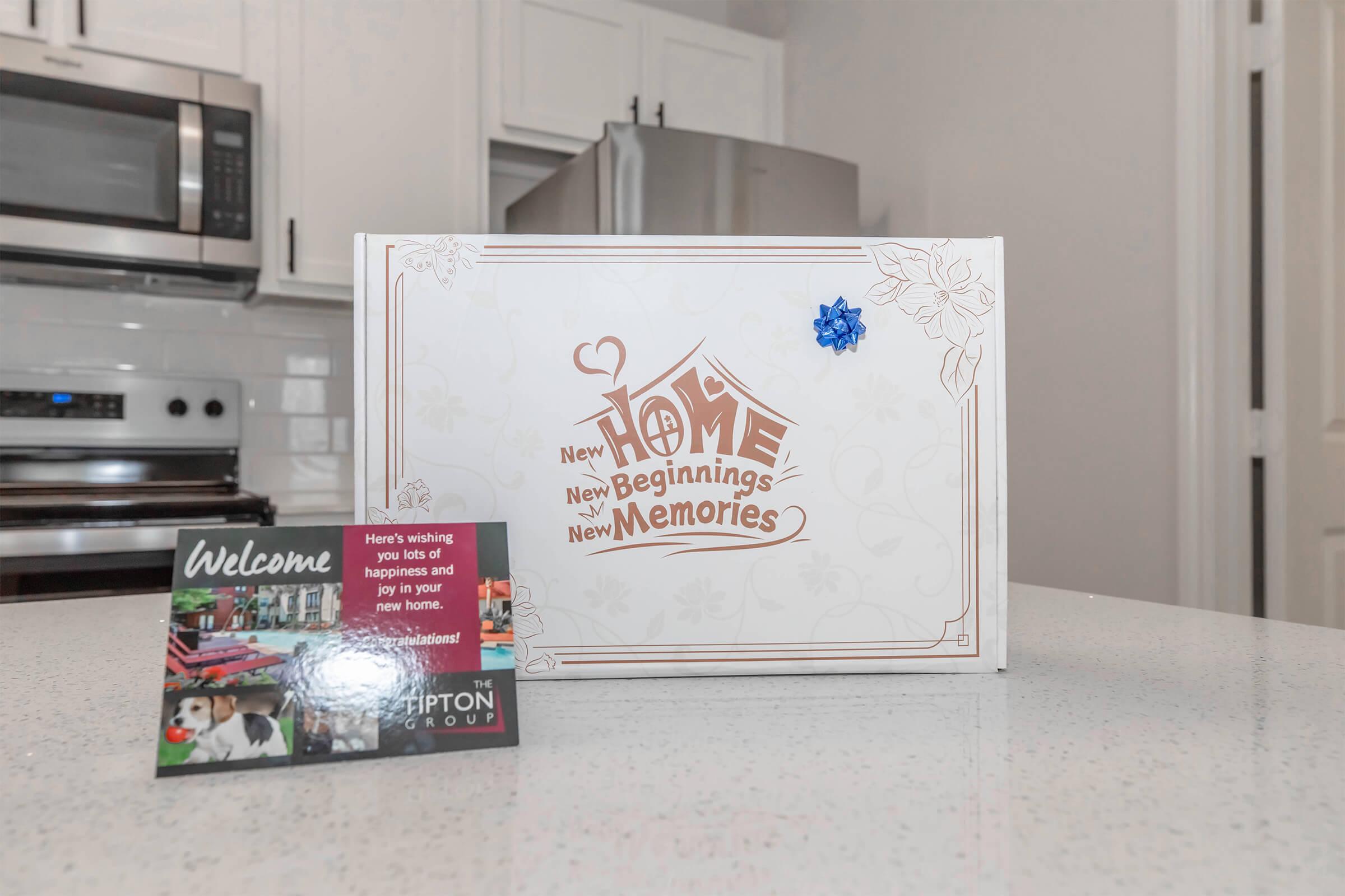 A decorative box labeled "New Home, New Beginnings, New Memories," featuring a blue flower embellishment, placed on a kitchen countertop. Next to it is a welcome card from a real estate group, wishing happiness and joy in the new home. The kitchen has modern appliances and a clean, bright aesthetic.