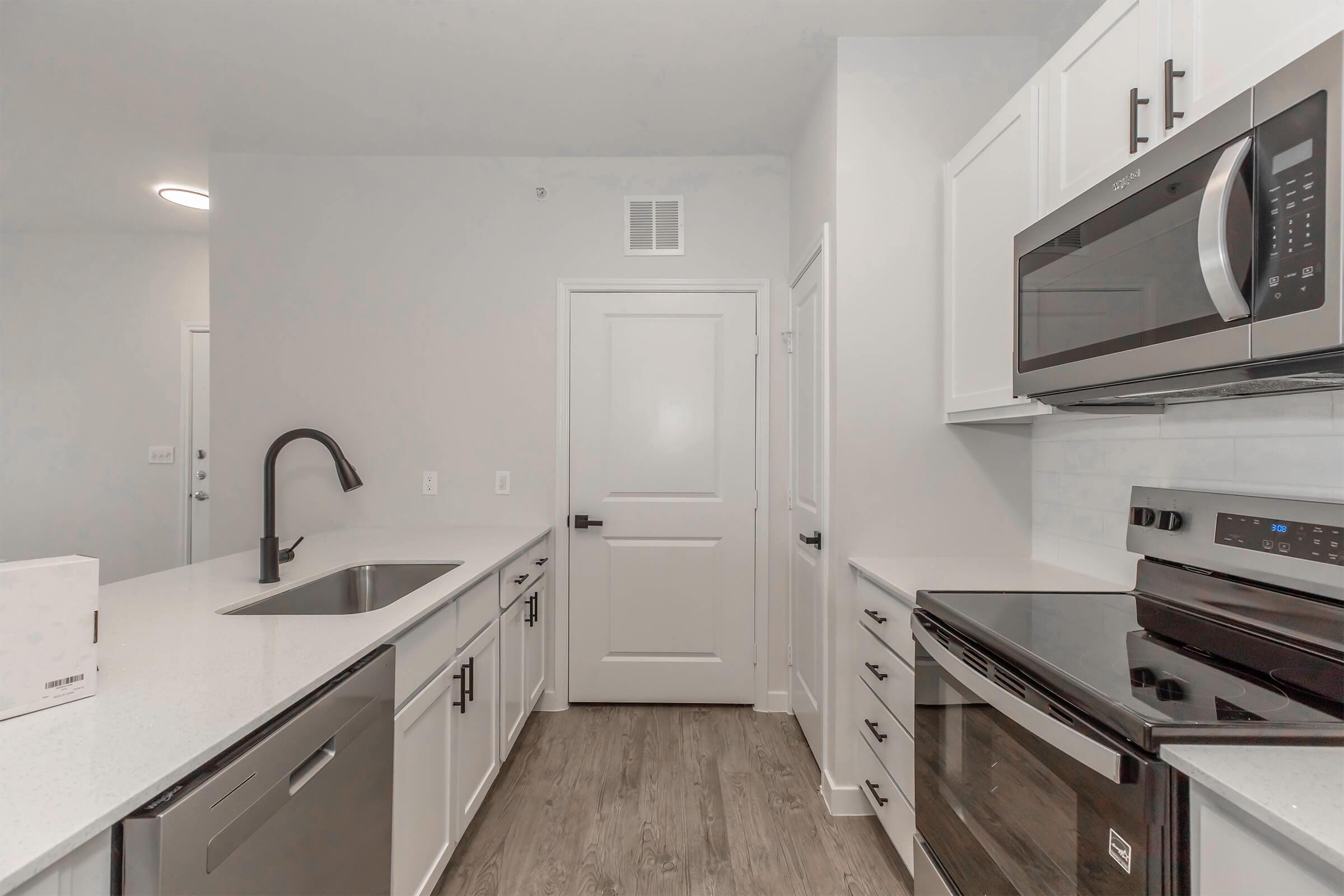 Modern kitchen featuring white cabinetry, stainless steel appliances including a microwave and oven, a single basin sink, and a sleek countertop. The space has light-colored walls and a wood-style floor, with a door leading to another room. Natural light illuminates the area.