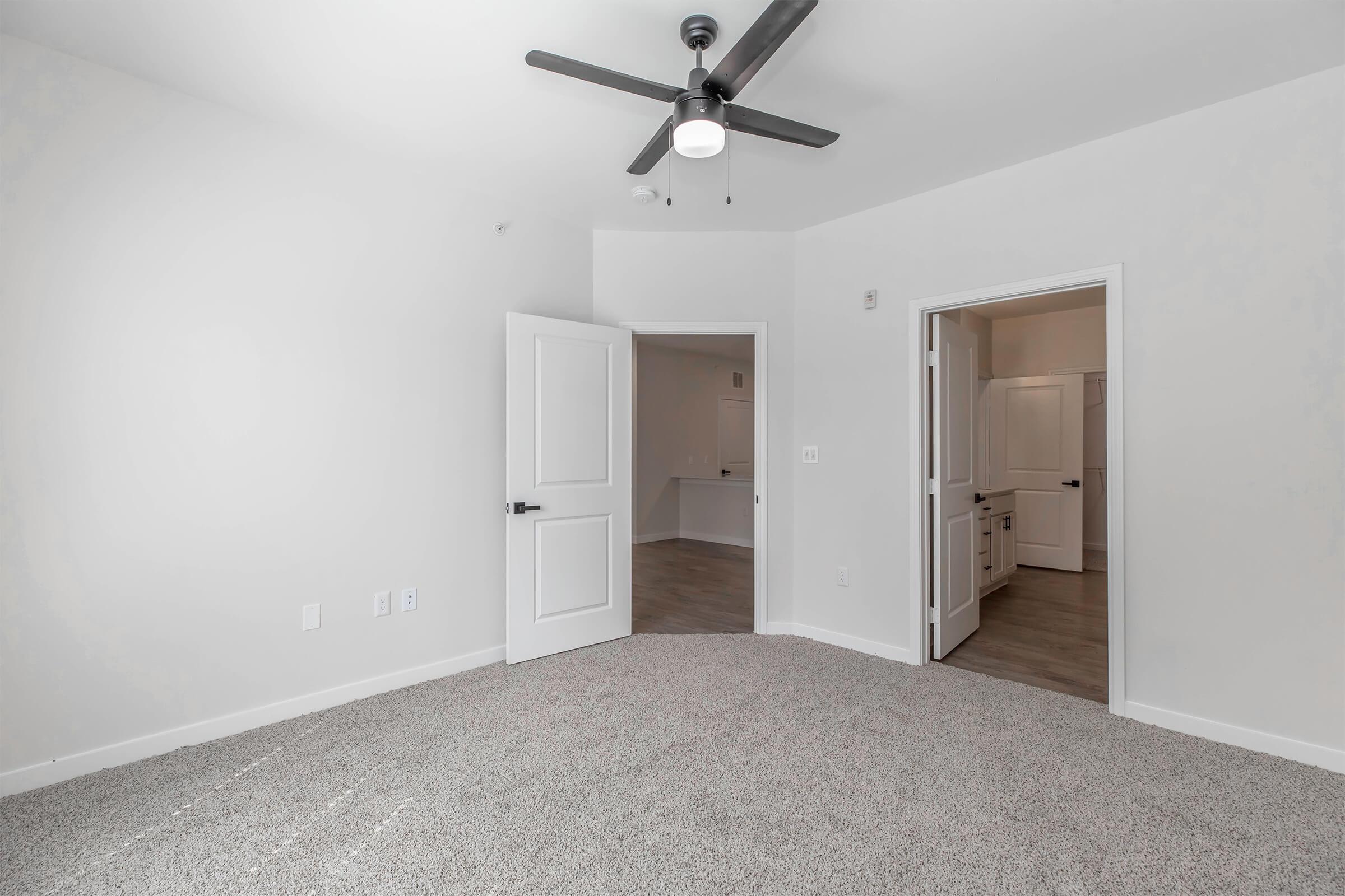 A spacious, empty room with light-colored walls and a ceiling fan. The floor is carpeted in a neutral tone, and there are two doorways leading to adjacent areas. Natural light fills the space, creating an inviting atmosphere.