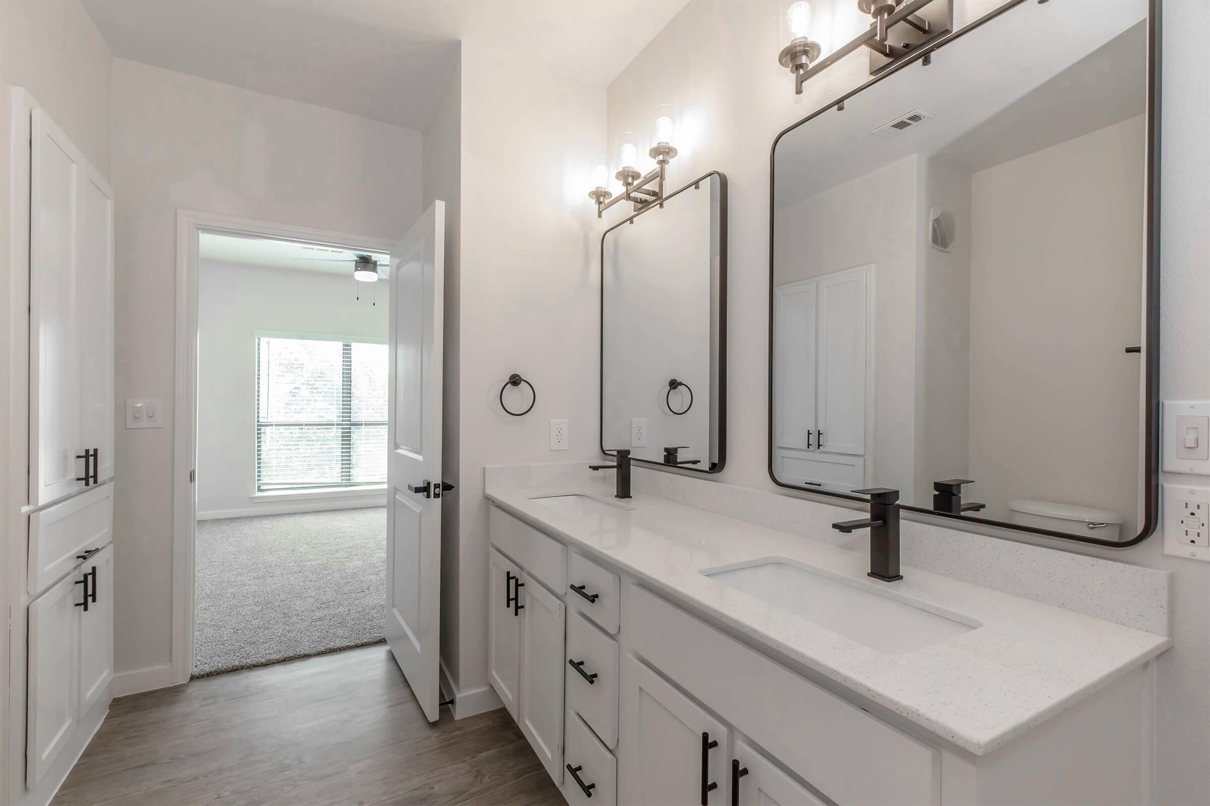 Modern bathroom featuring a double sink vanity with white cabinetry, sleek black faucets, and large rectangular mirrors. The walls are painted a light color, and the flooring is a warm wood tone. A doorway at the back leads to a well-lit room with windows, suggesting a connected living space.
