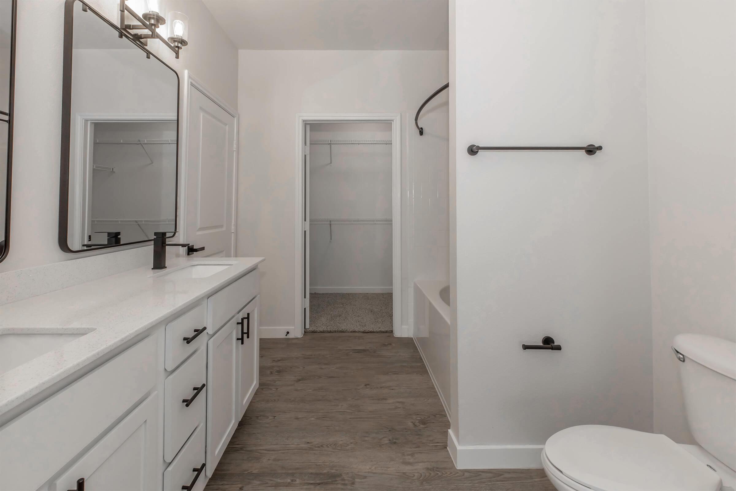 A modern bathroom featuring a double sink vanity with black fixtures, a large mirror, and a seamless design. The floor is finished with wood-like tiles, and there is a white bathtub with a shower. A closet is visible in the background, emphasizing a spacious and clean aesthetic.