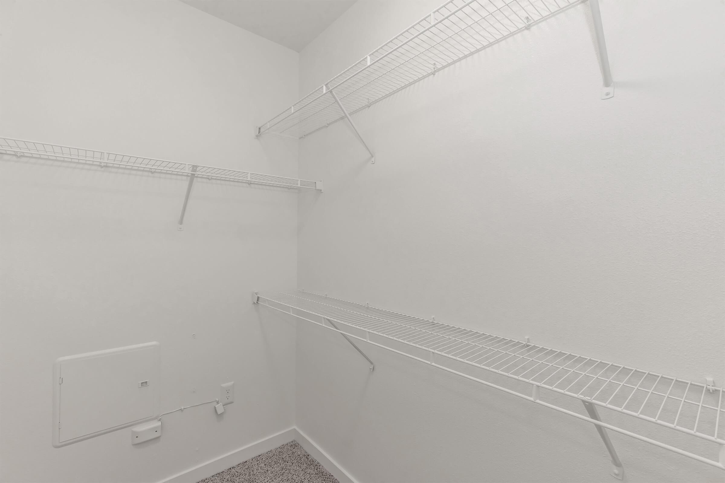 Empty closet featuring white wire shelving mounted on the walls. The walls and ceiling are painted white, and the floor is covered in a gray carpet. There are no items stored in the closet, providing a clean and minimalistic appearance.
