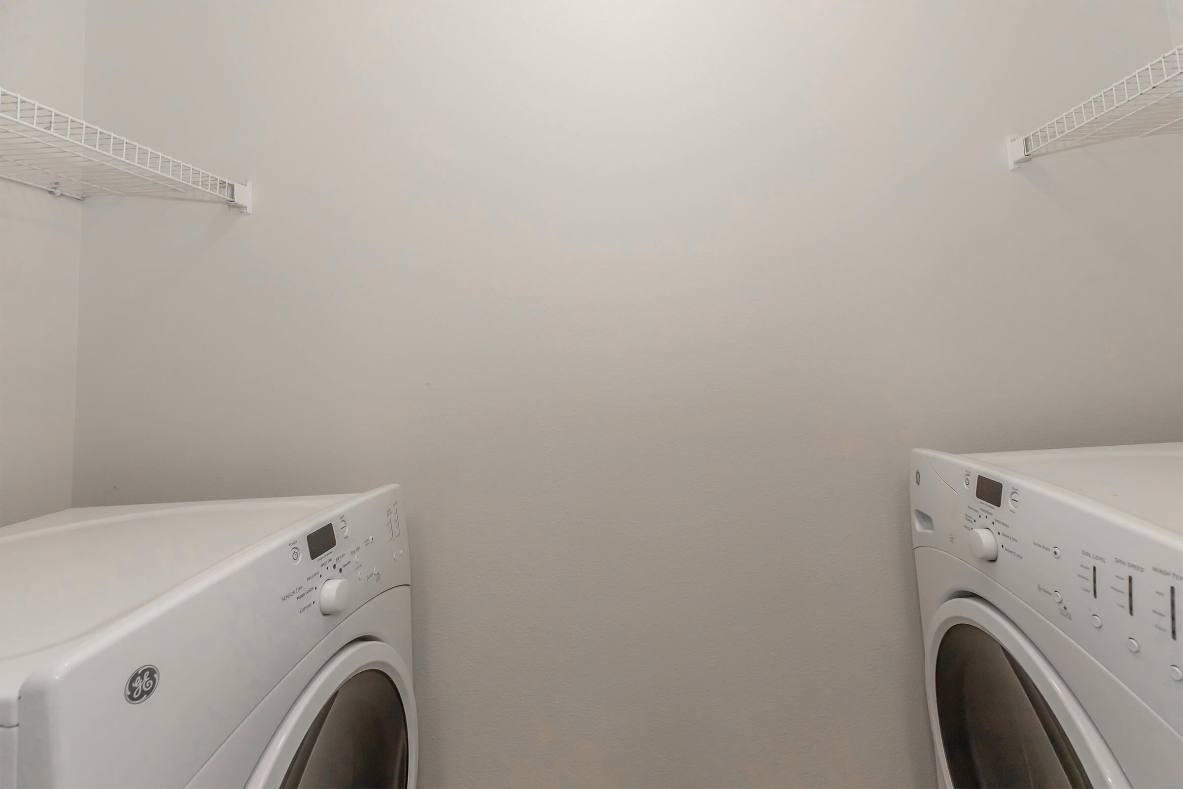 A spacious laundry room featuring two white appliances side by side. The washing machine and dryer have sleek designs, and there are wire shelves above them for storage. The walls are plain and light-colored, providing a clean and organized look.